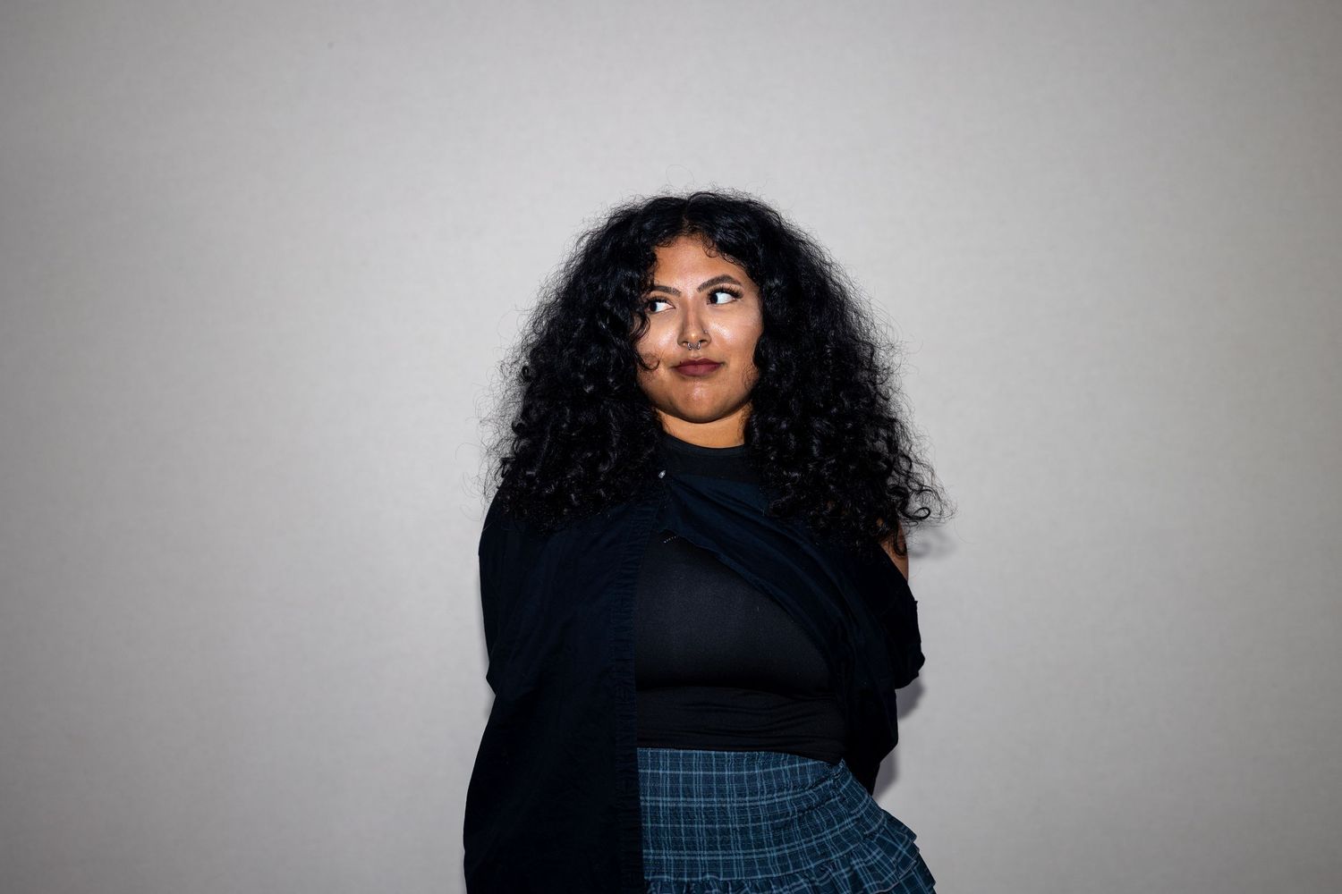 A plus-size woman with curly hair in a black top and plaid skirt looks upward against a gray background.