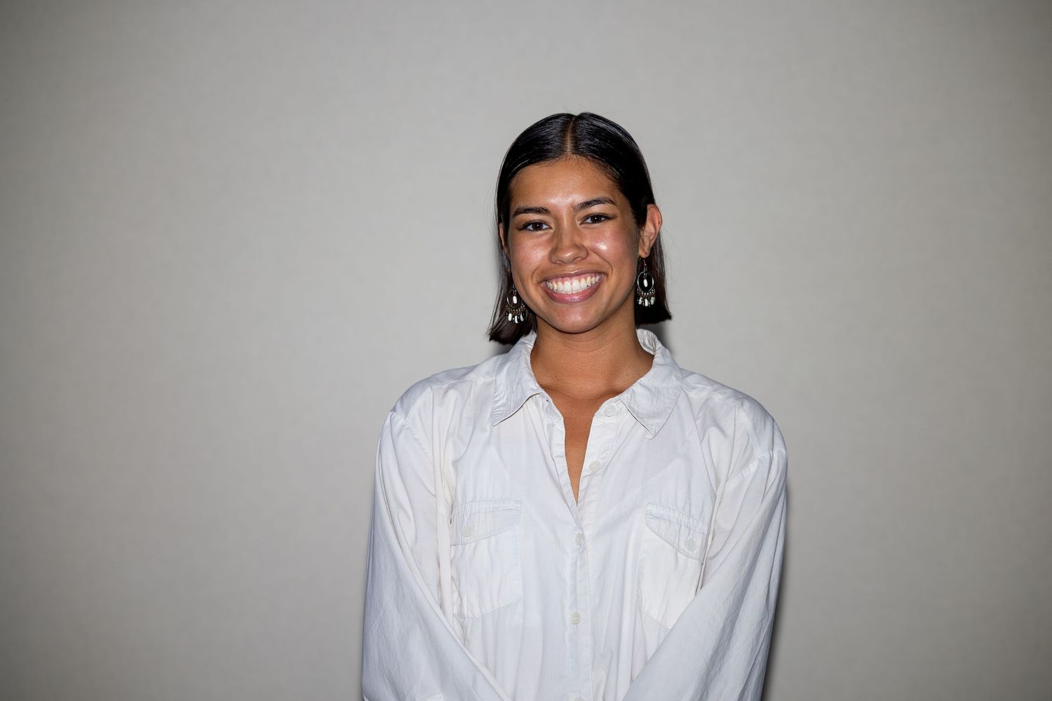 Smiling woman in white button-up shirt with dangling earrings poses against neutral background.