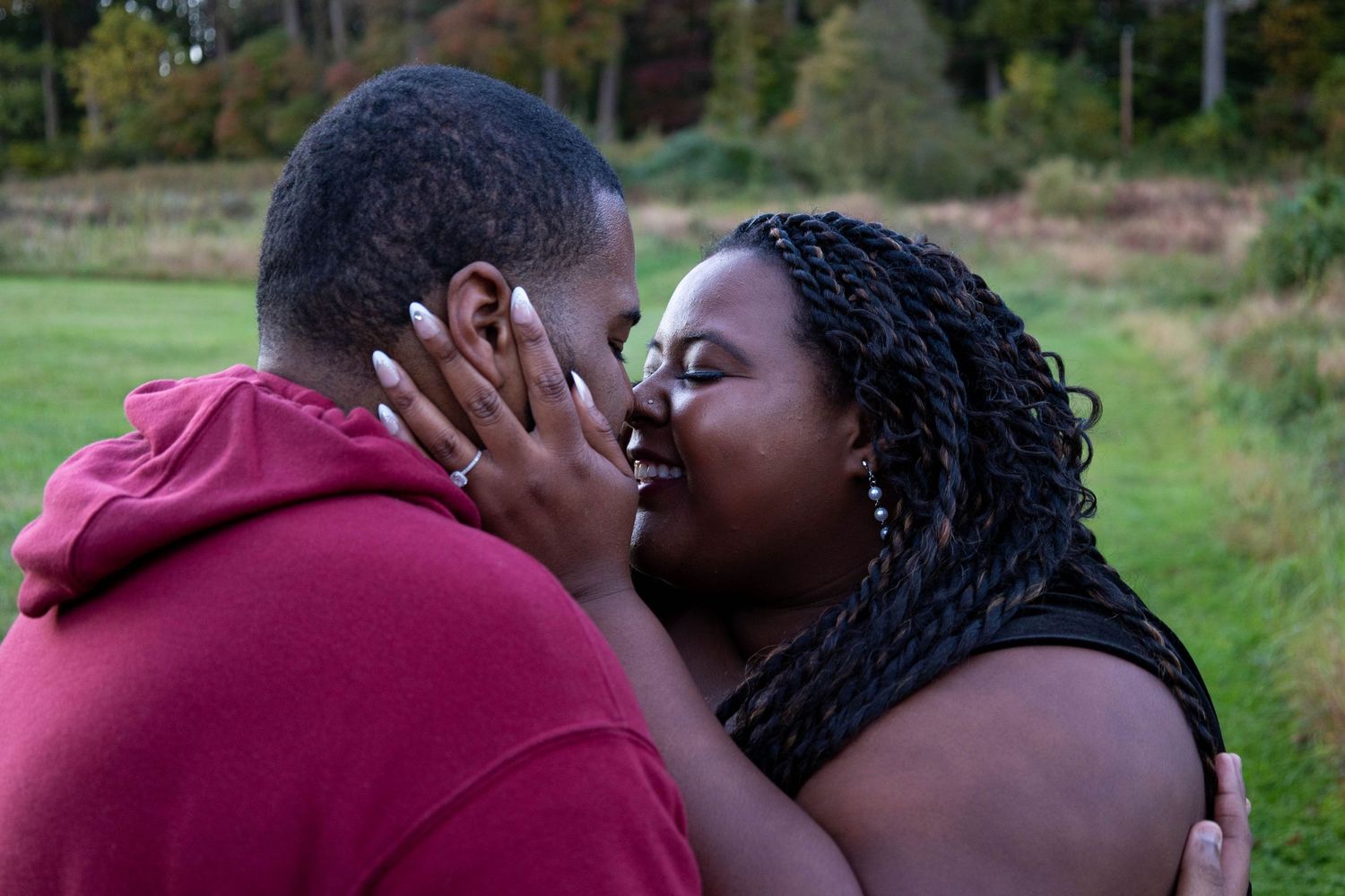 A loving Black couple shares a kiss outdoors, the woman displaying an engagement ring on her hand.