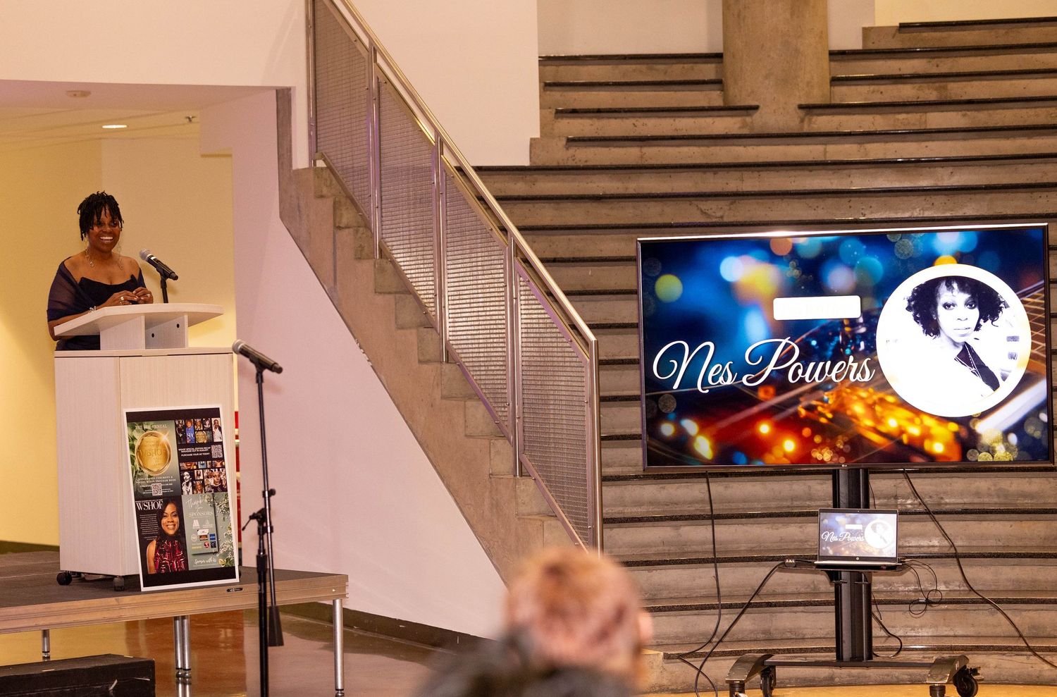 Woman speaking at podium beside a screen displaying Nes Powers tribute at a formal event.