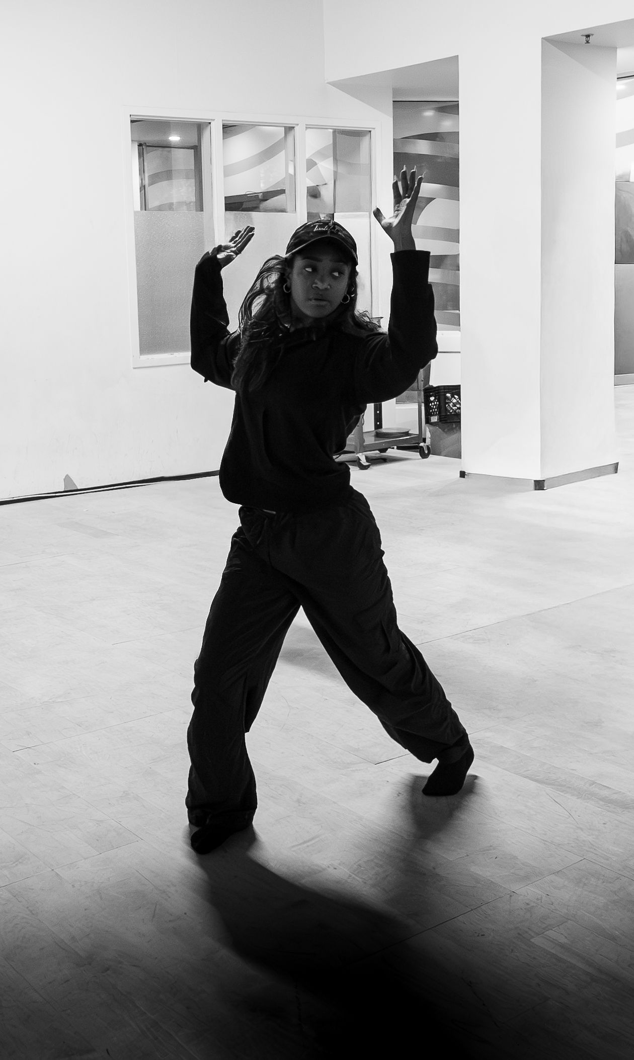 Black and white photo of a dancer in a cap and baggy clothes striking a dynamic hip-hop pose in a studio.