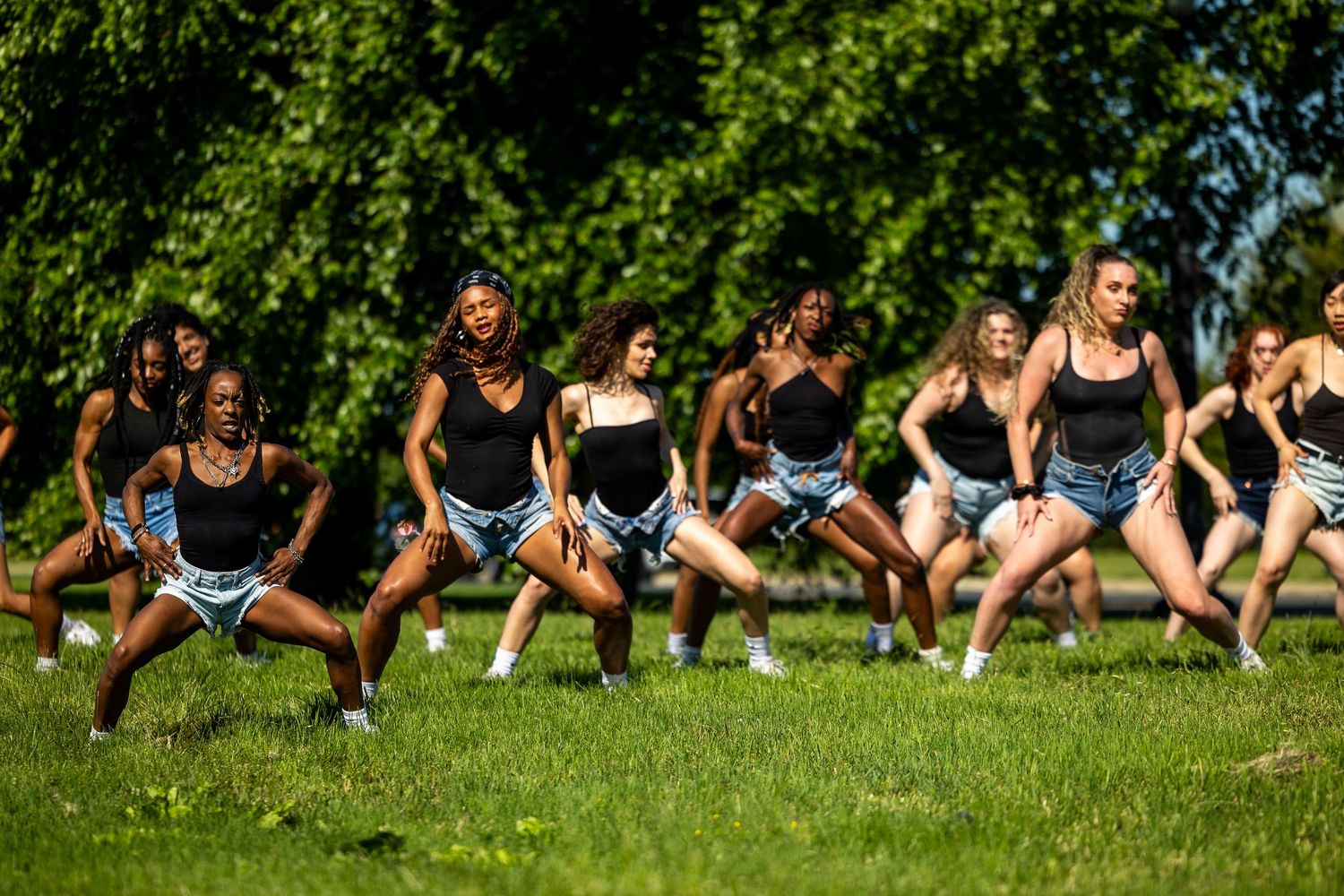 Group of young women in black tops and denim shorts performing a synchronized dance routine outdoors on a sunny day.