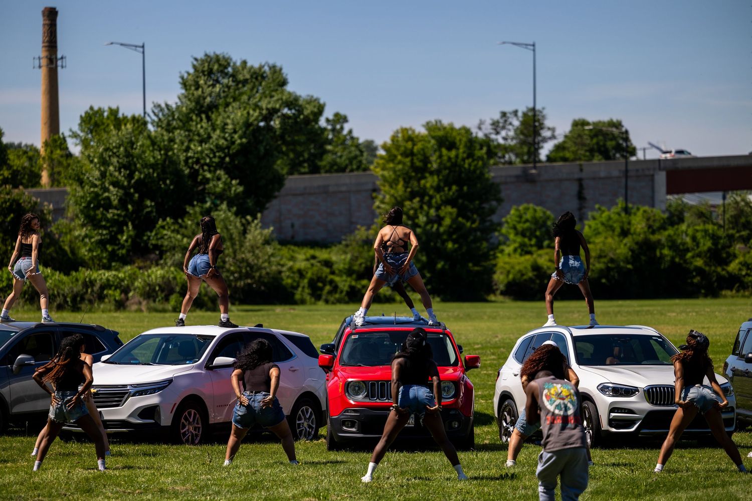 Cheerleaders performing stunts on cars in a grassy field with trees and a smokestack in the background.