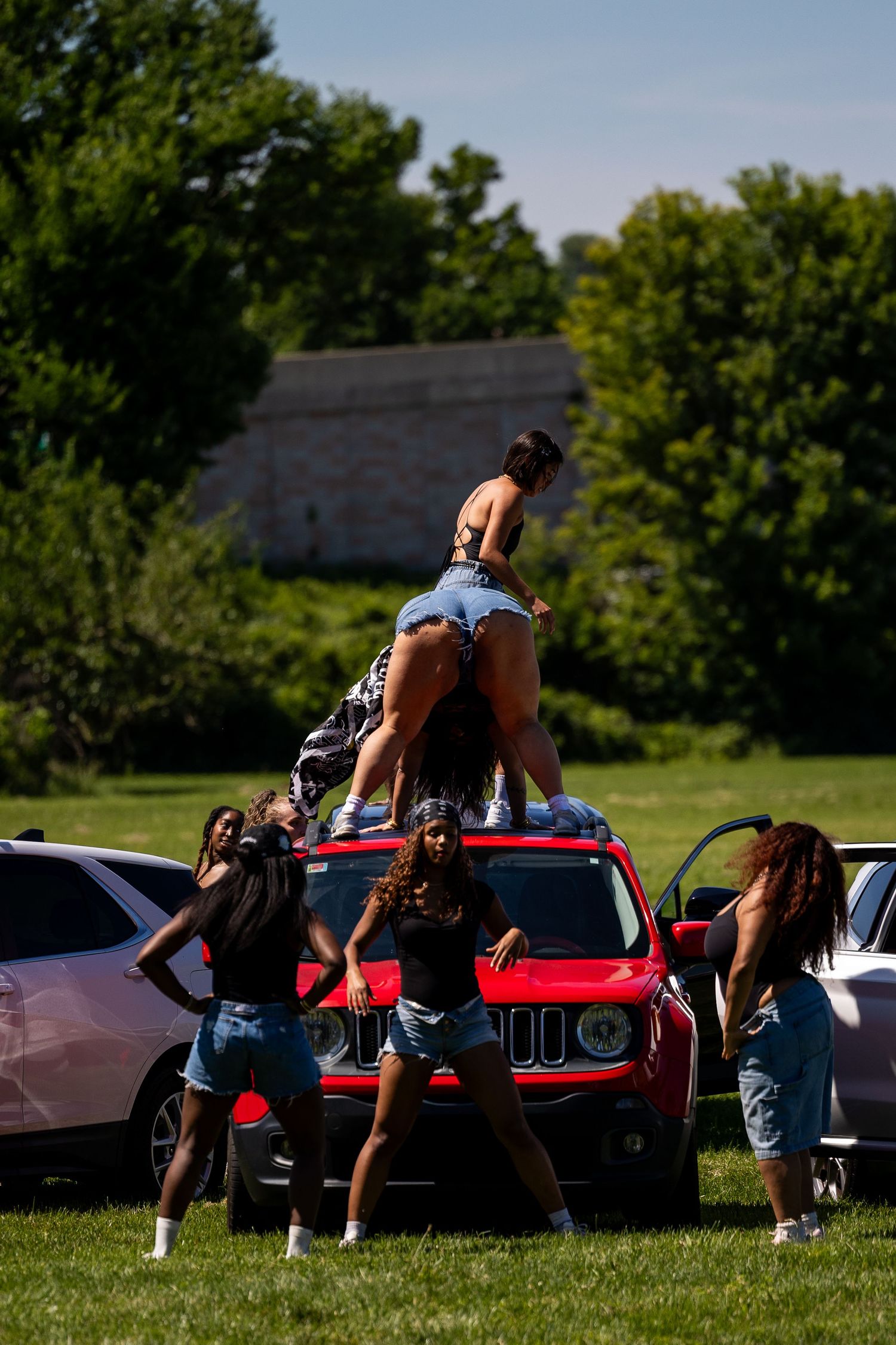 Group of young women dancing around a red Jeep outdoors on a sunny day with green trees in the background.