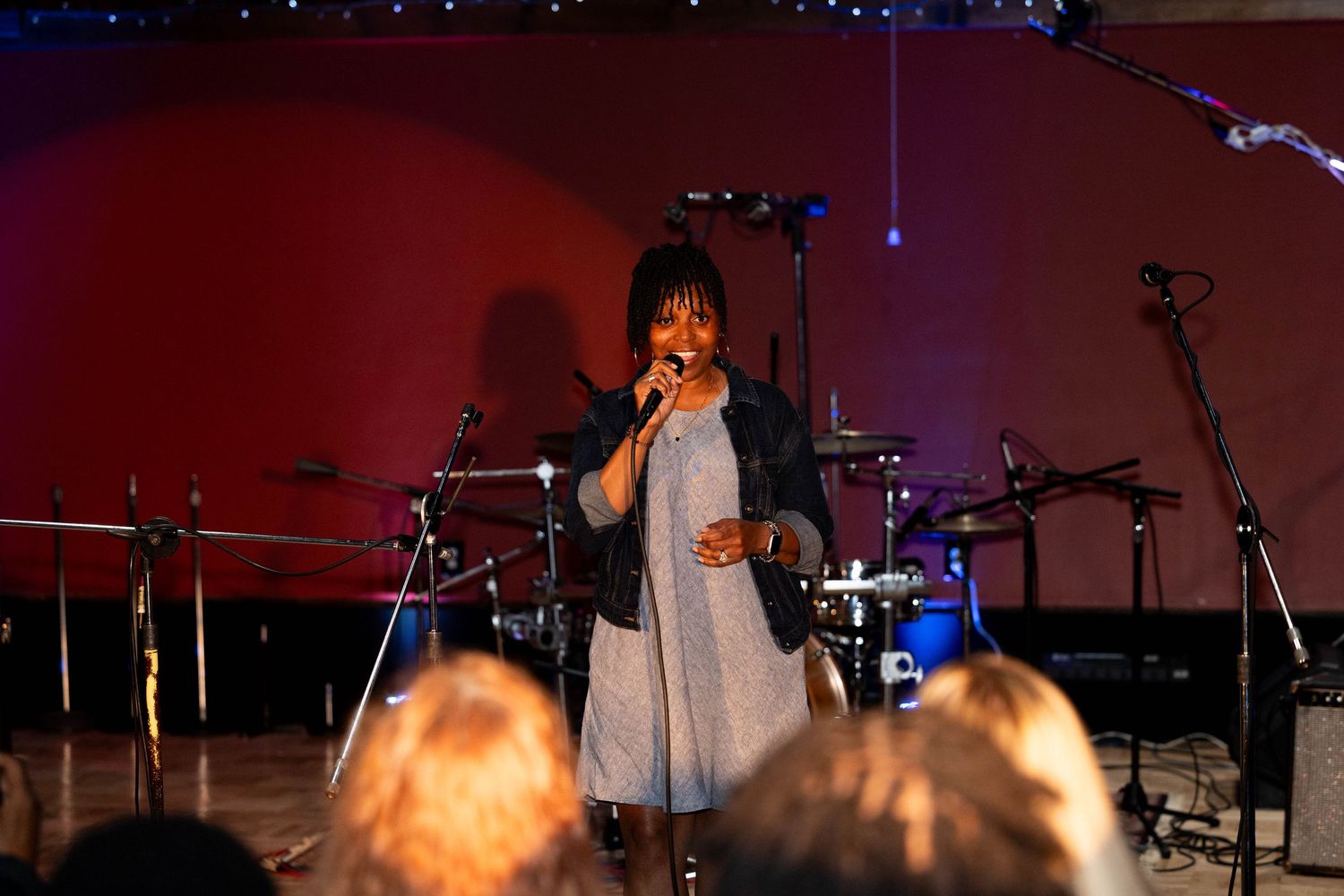 A woman singer performs on stage holding a microphone with drums and red lighting behind her.