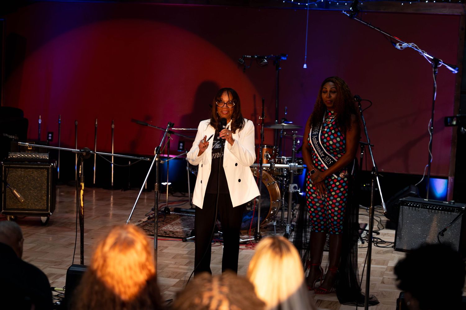 Two women performing on stage, one in a white blazer speaking into a microphone at a live music event.