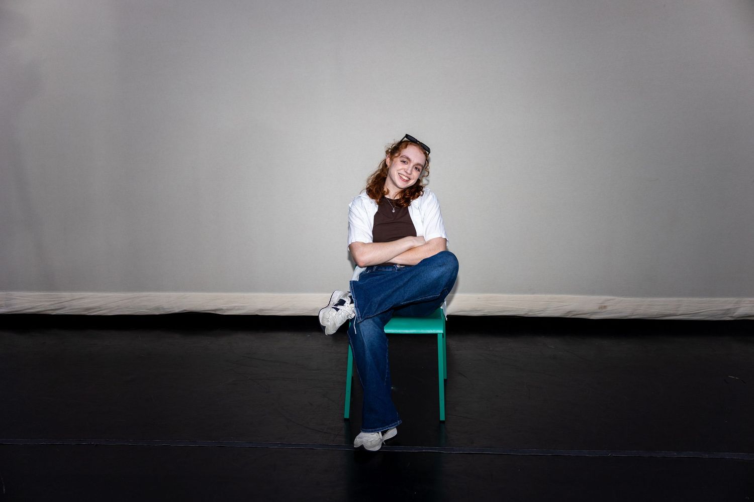 A young woman in a brown top, jeans, and white sneakers sits relaxed on a green chair in a studio.