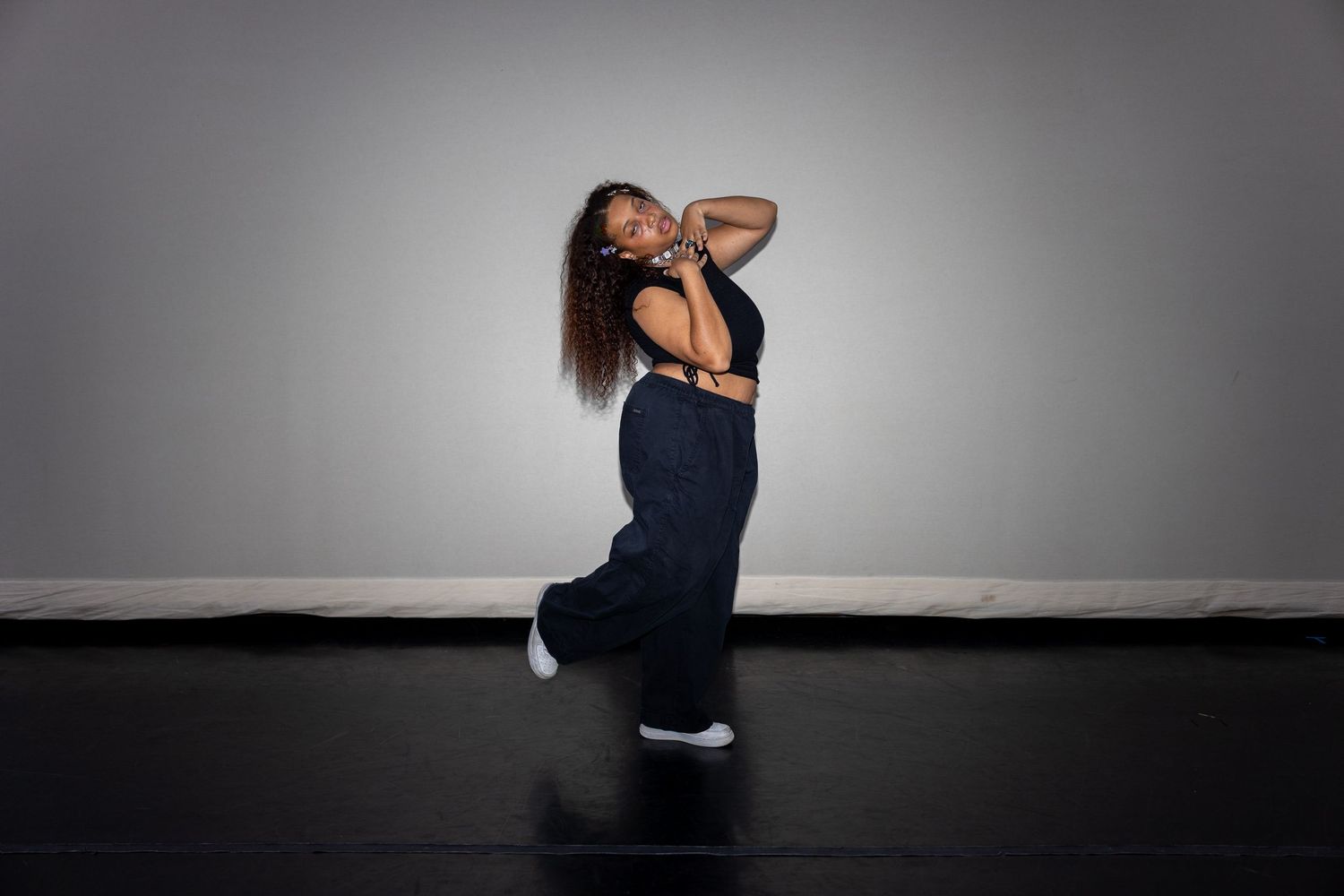 A dancer in a black crop top and baggy pants poses gracefully with hand behind head in a studio.