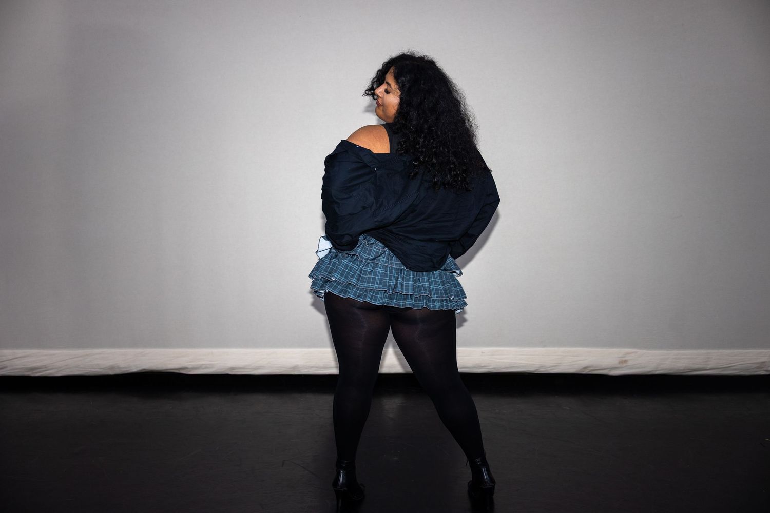 Curvy woman in denim jacket and plaid mini skirt poses with back to camera against gray backdrop.