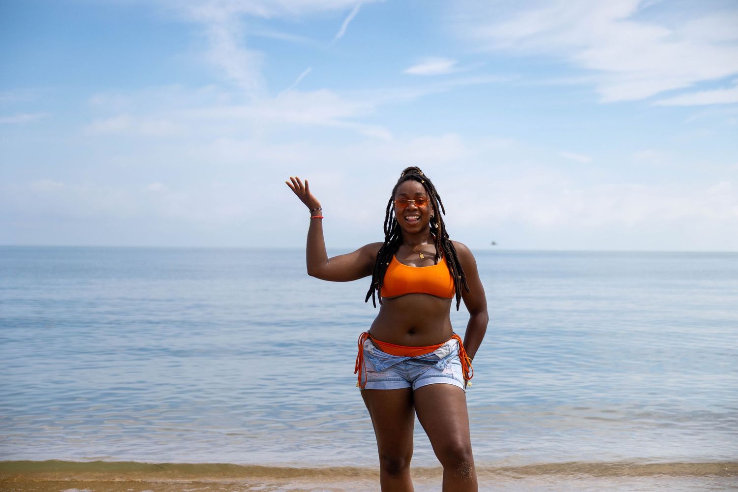 Happy Black woman in orange bikini top and denim shorts waving at beach with blue ocean and sky.