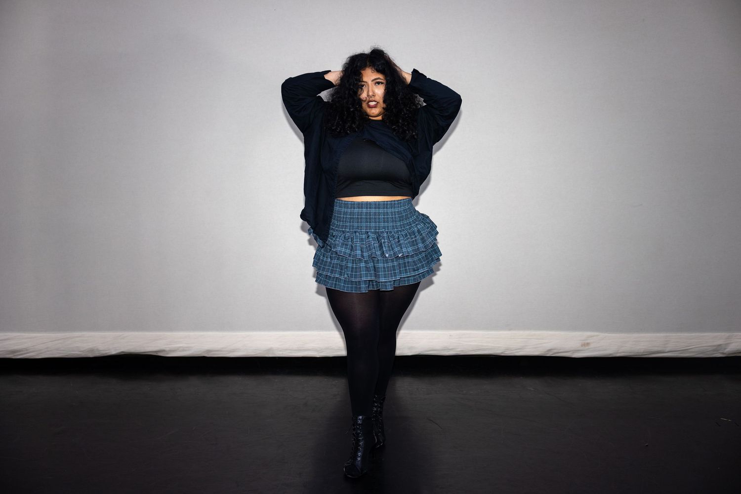 Plus-size woman posing in black crop sweater and plaid mini skirt with tights in a dark studio.
