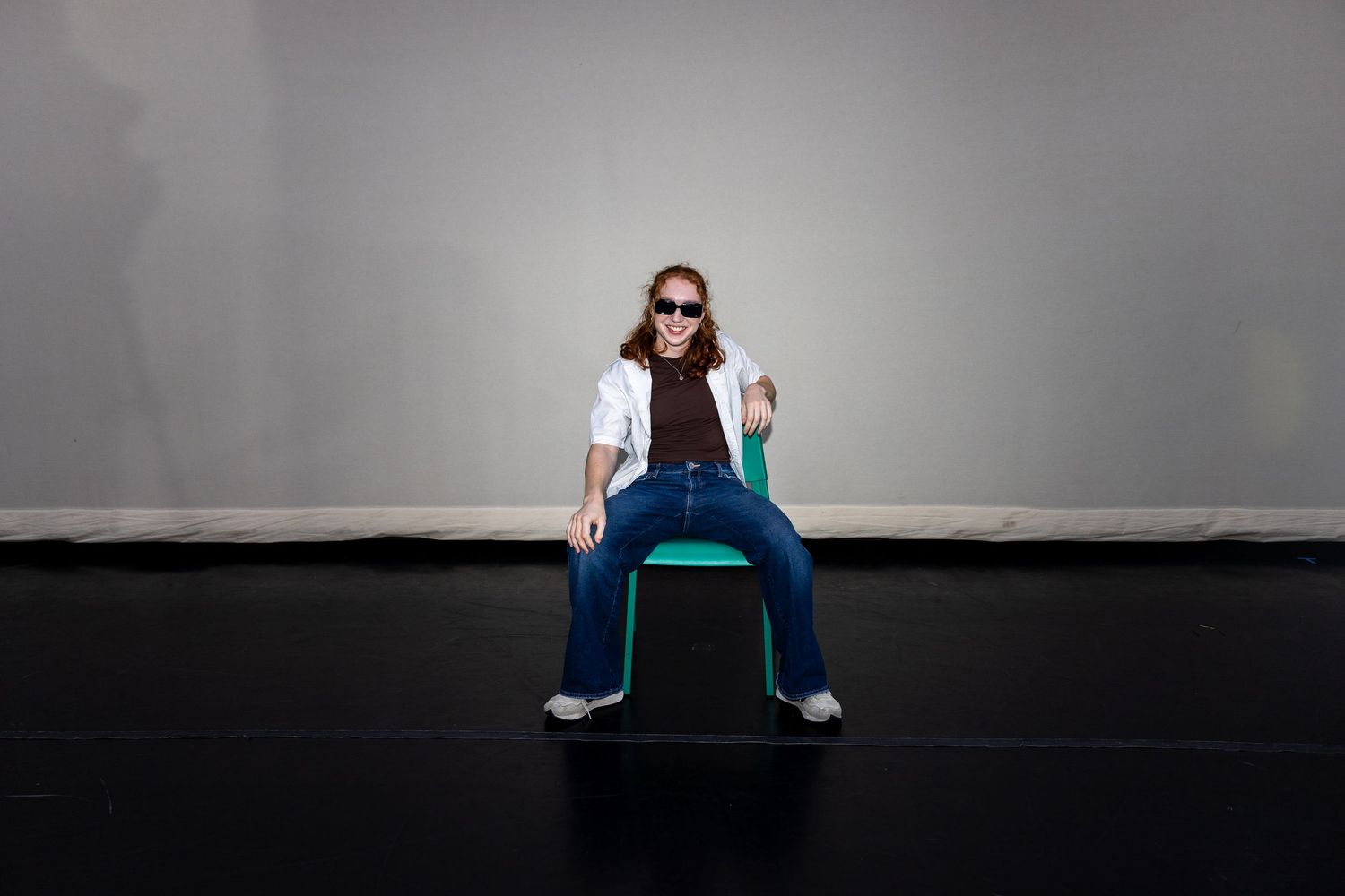 A dancer in sunglasses and jeans sits confidently on a green chair on a black stage.