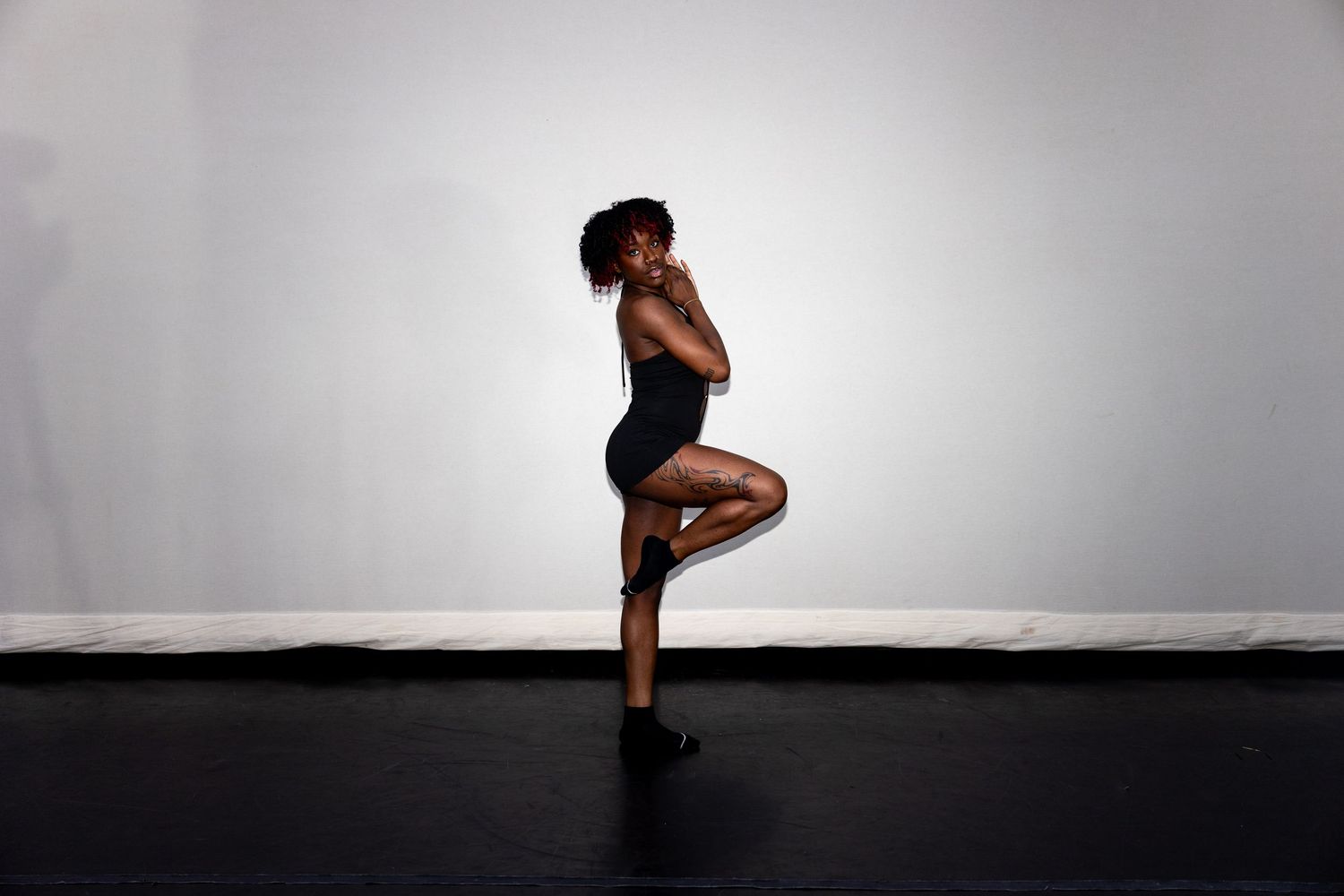 Athletic dancer balances on one leg in black bodysuit against white studio wall on dark floor.