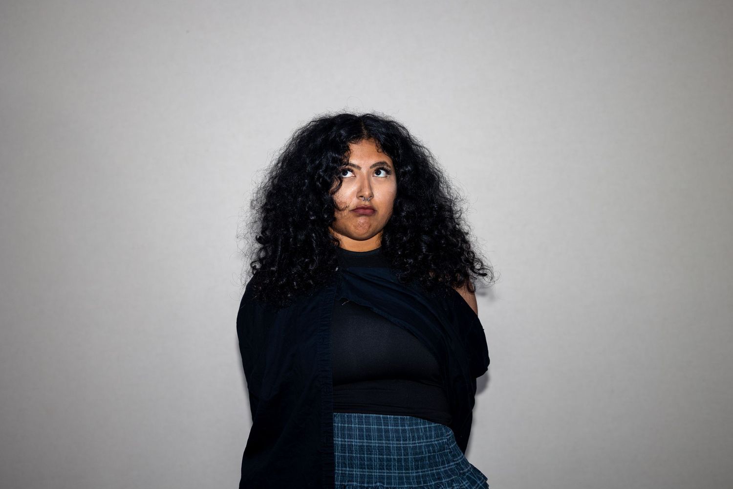 Young woman with voluminous curly hair wearing black top and blue plaid skirt against gray wall.