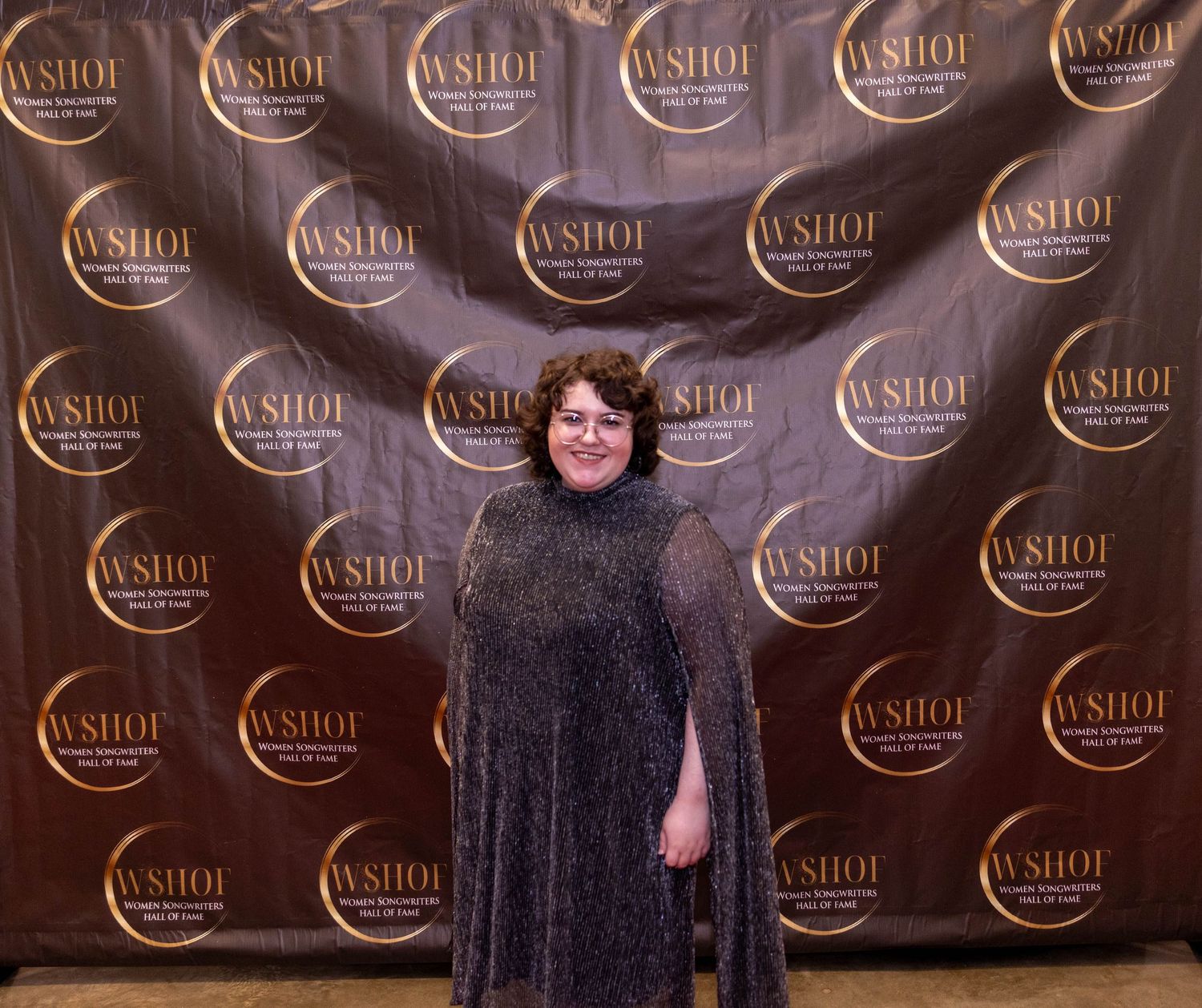 Woman in sparkly dress posing at WSHOI event backdrop in elegant black and gold step-and-repeat banner.