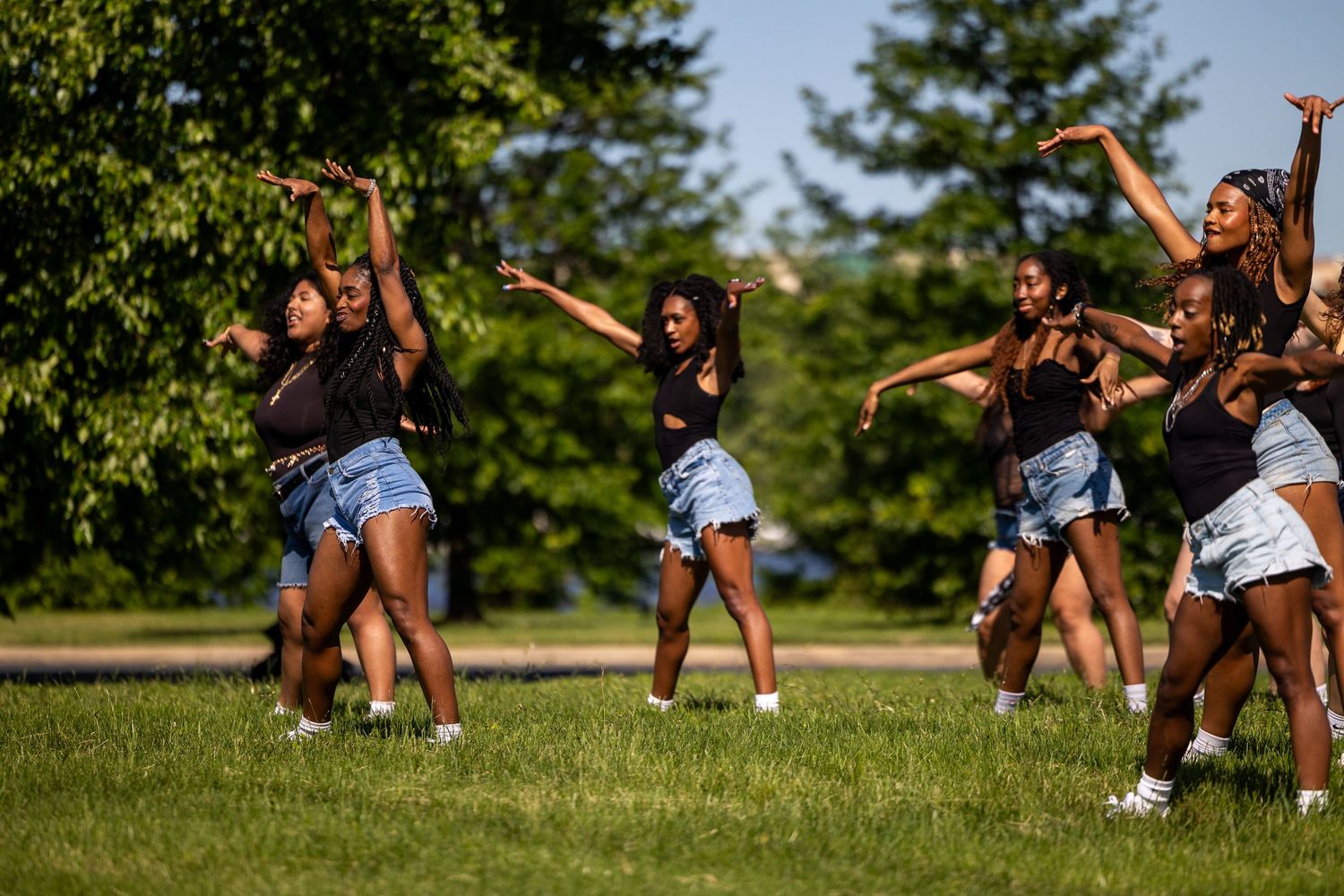 Group of young women in black tops and denim shorts performing a synchronized dance routine outdoors on green grass.