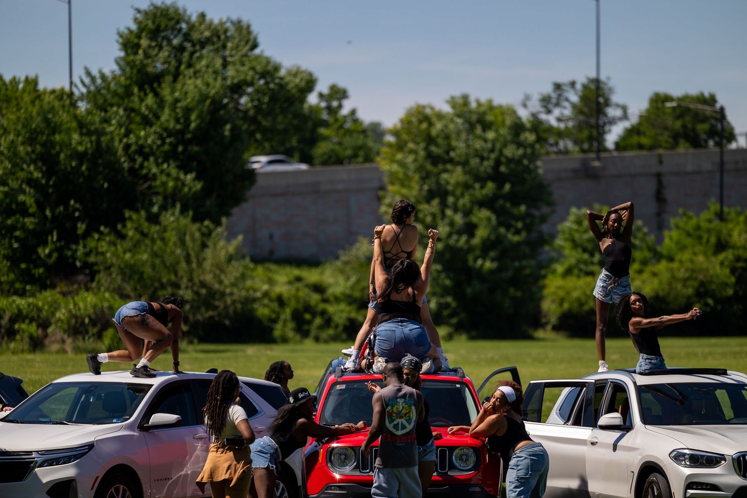 People dancing on top of cars at an outdoor gathering on a sunny day with green trees in the background.