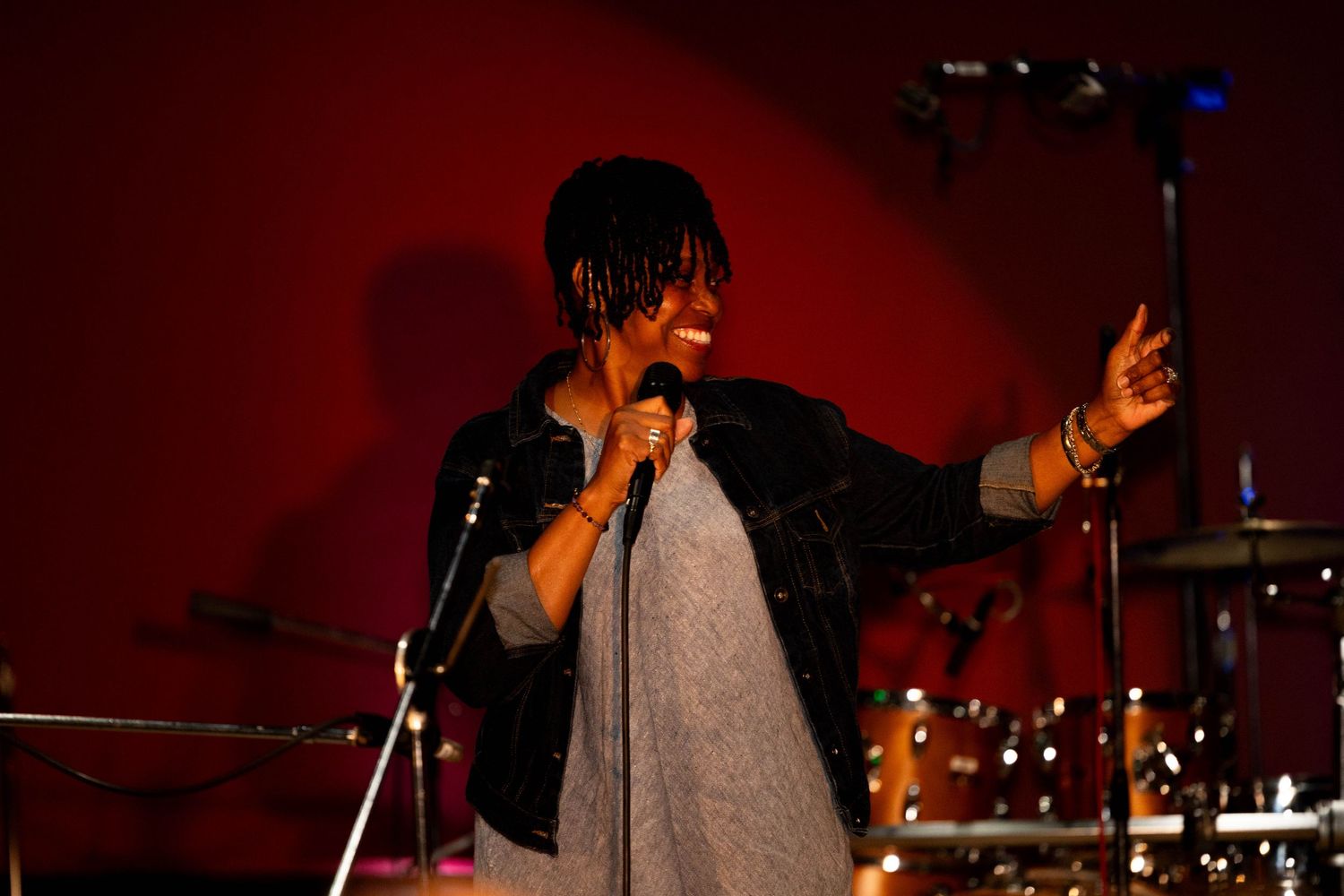 Woman singer performing on stage holding microphone, wearing gray dress and black jacket, red wall background.