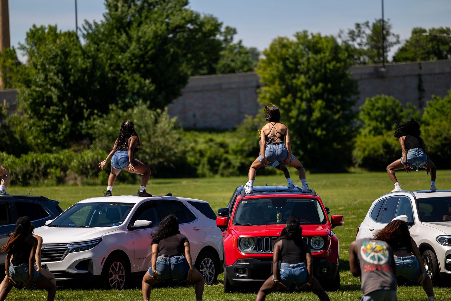 Dancers perform on top of cars in a sunny outdoor lot, with others doing handstands against vehicles on green grass.
