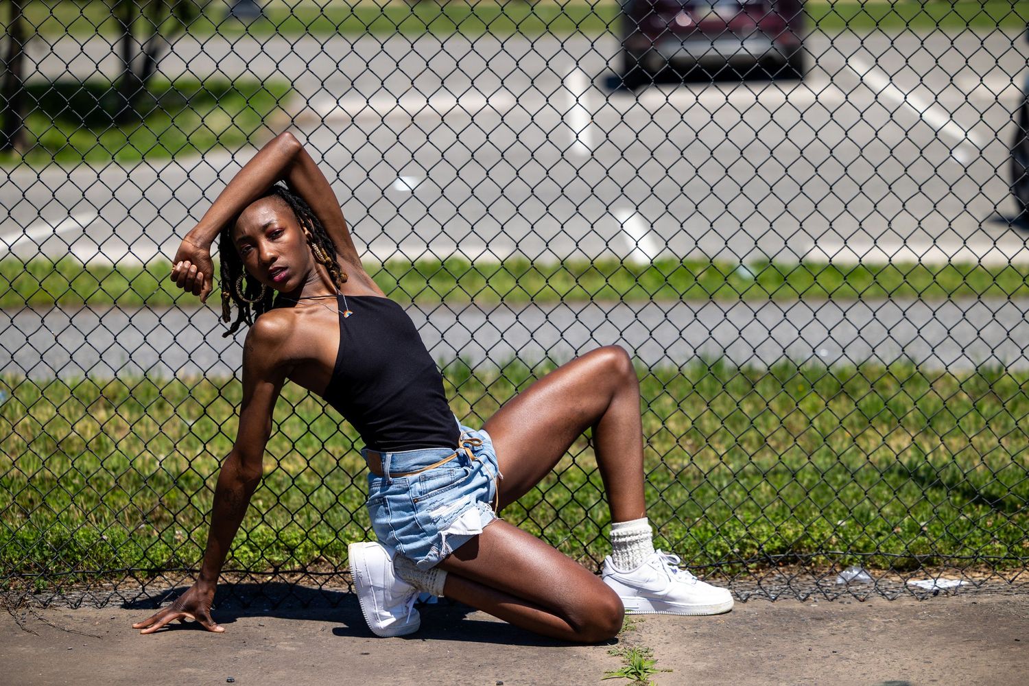 Young woman in black top and denim shorts posing confidently by a chain-link fence on a sunny day.