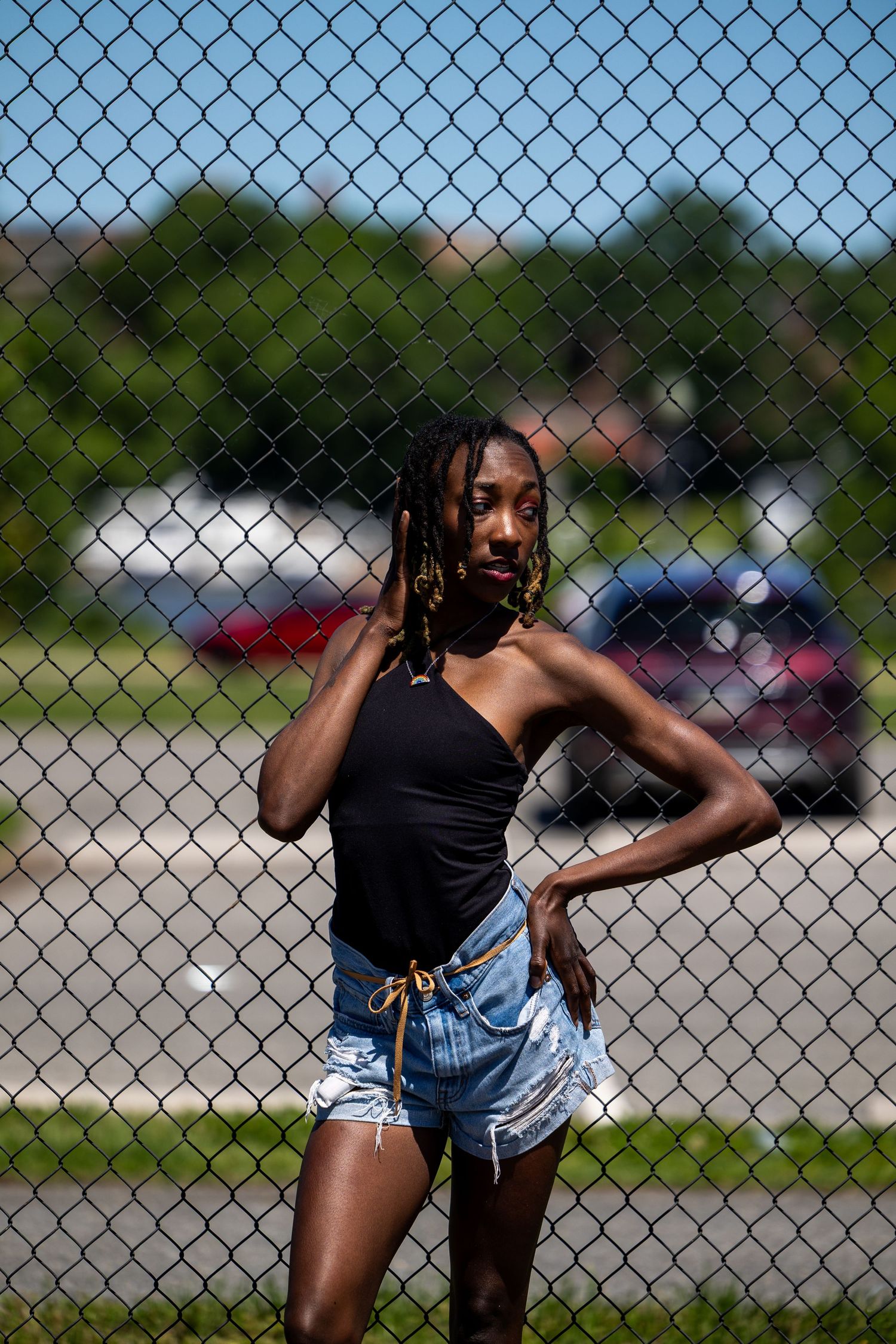 Stylish young Black woman with braids in black one-shoulder top and denim shorts leaning against chain-link fence.
