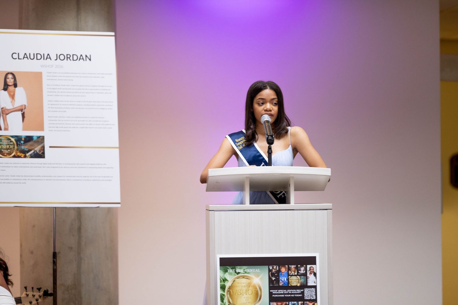 Young woman in white dress and sash speaks at podium near Claudia Jordan display at formal event.