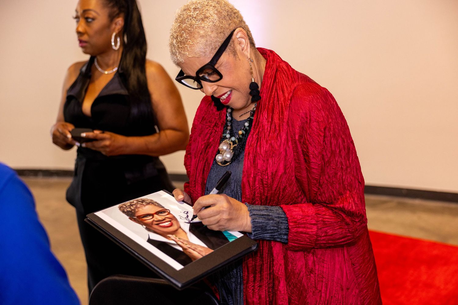 Elderly woman in red outfit signing a portrait artwork at a formal event.