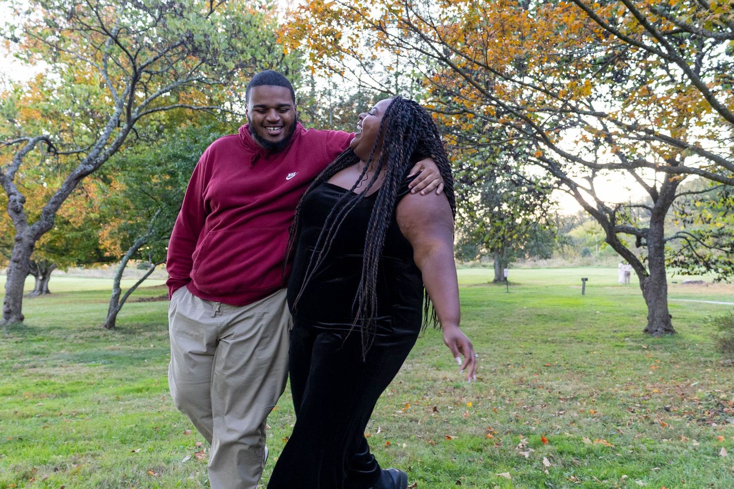 Two friends laughing and embracing outdoors in a park with autumn trees in the background.