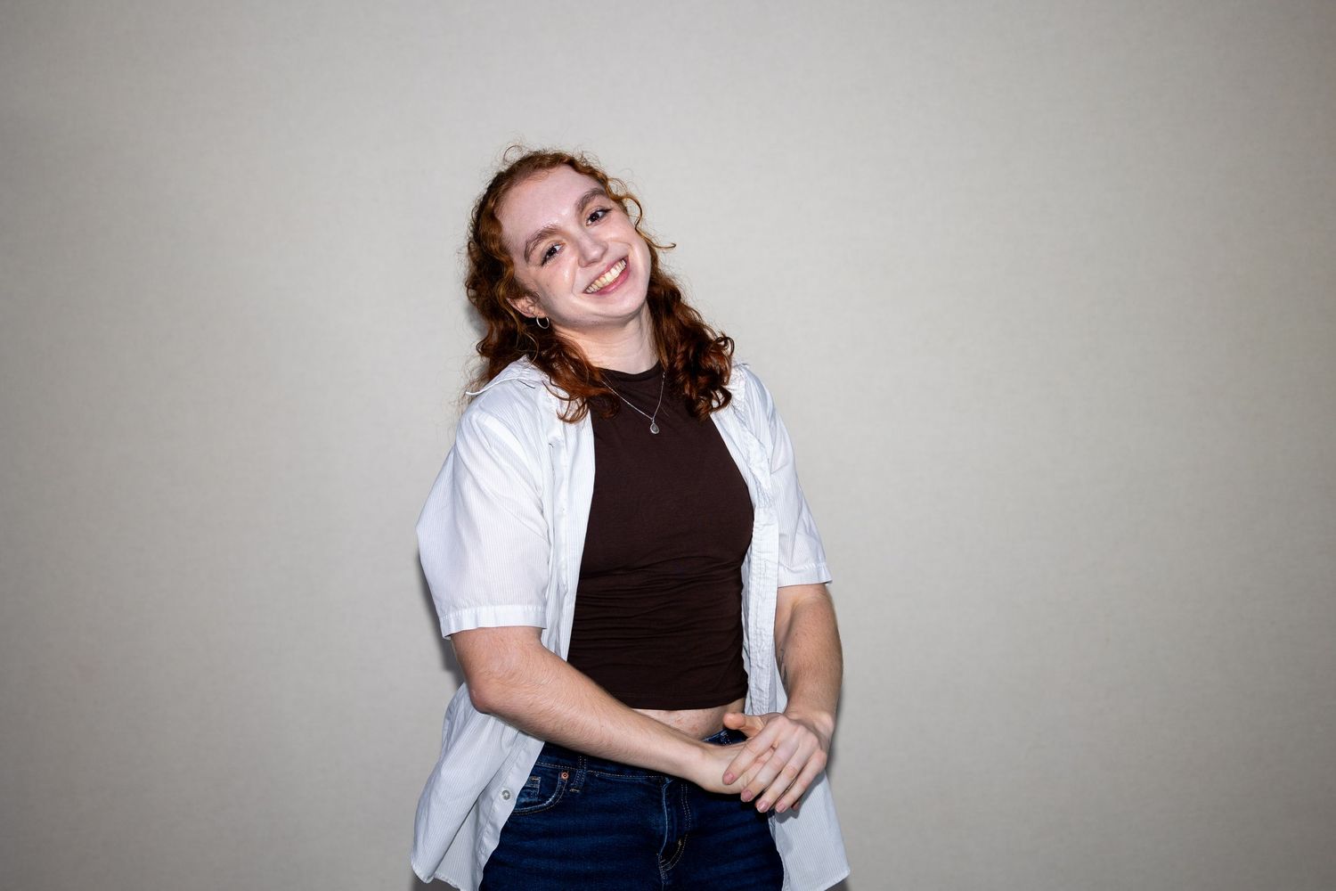 Smiling young woman with curly auburn hair wearing black top and white button shirt with blue jeans posing indoors.
