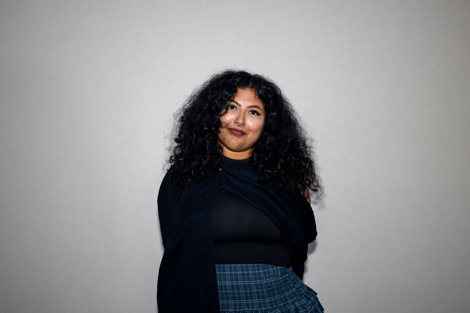 Young woman with voluminous curly hair wearing a black turtleneck and plaid skirt, smiling softly against a gray wall.