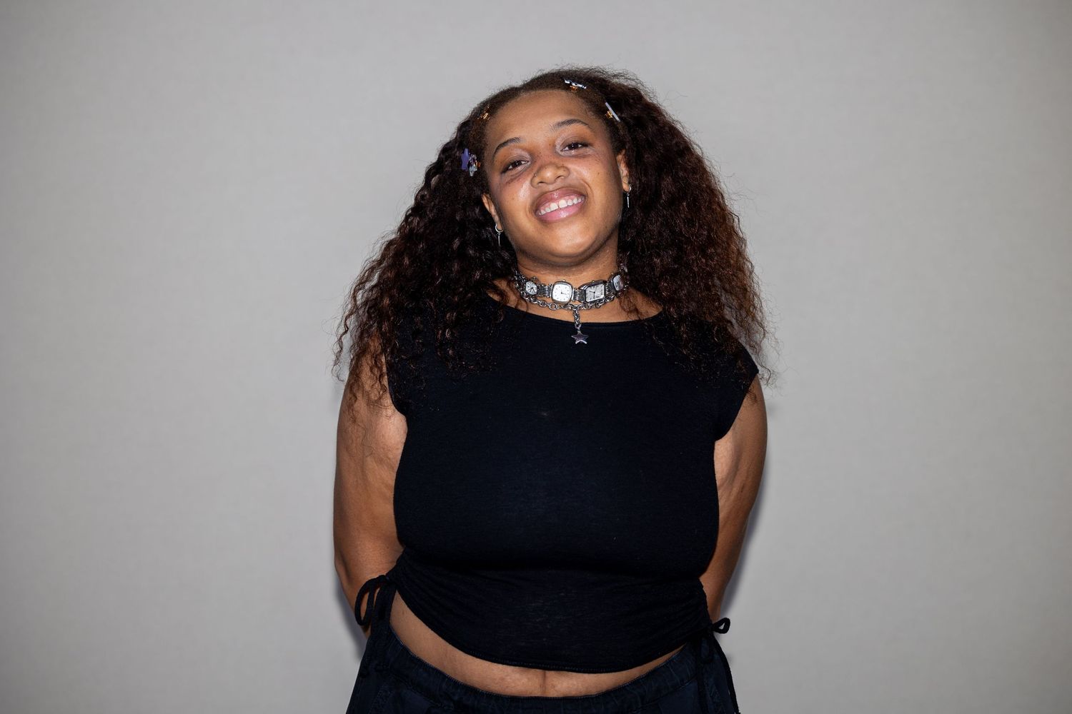 A young woman with curly hair wearing a black crop top and silver choker necklace smiles brightly.