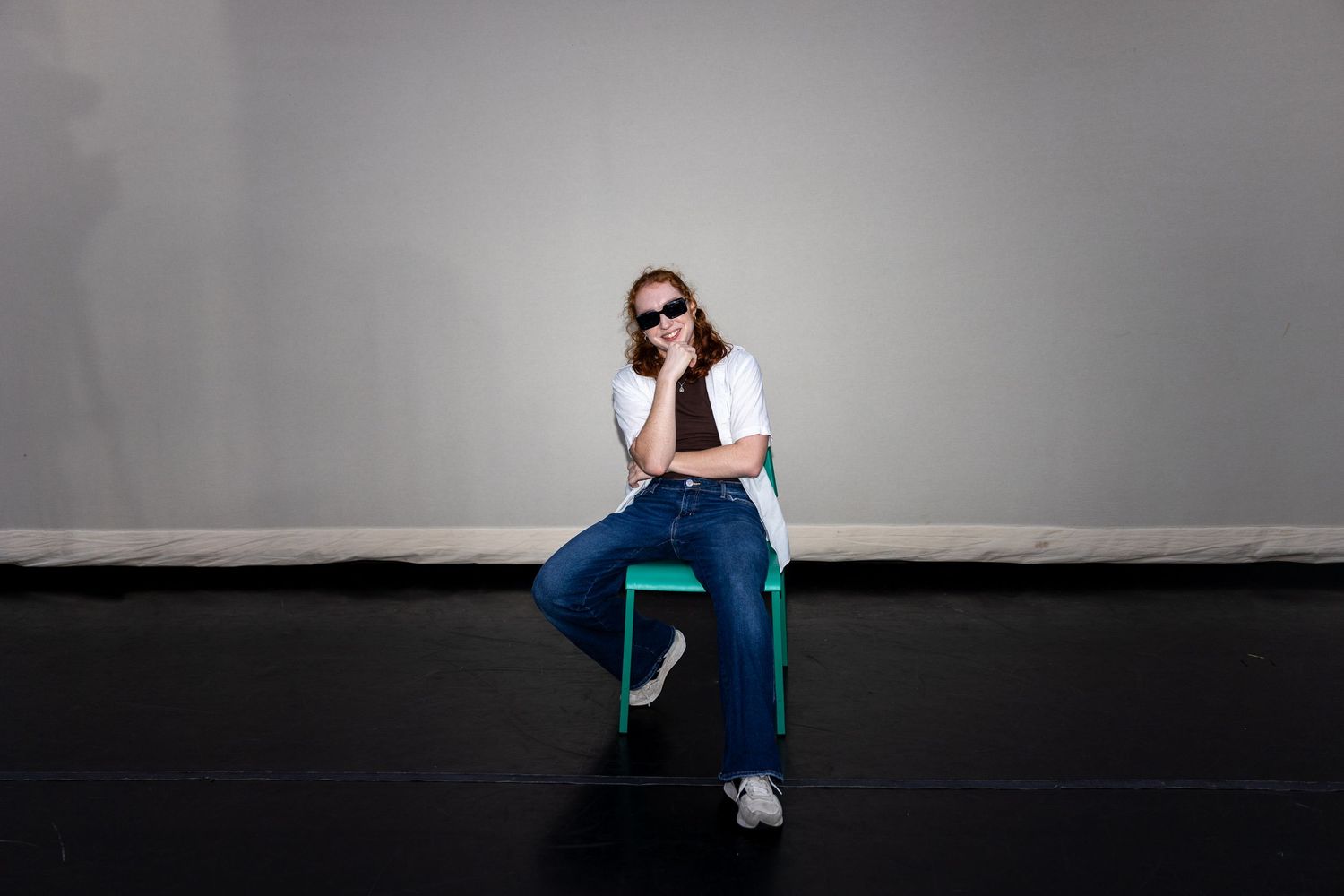 Woman in white shirt and jeans sitting on a teal chair wearing sunglasses on a stage.