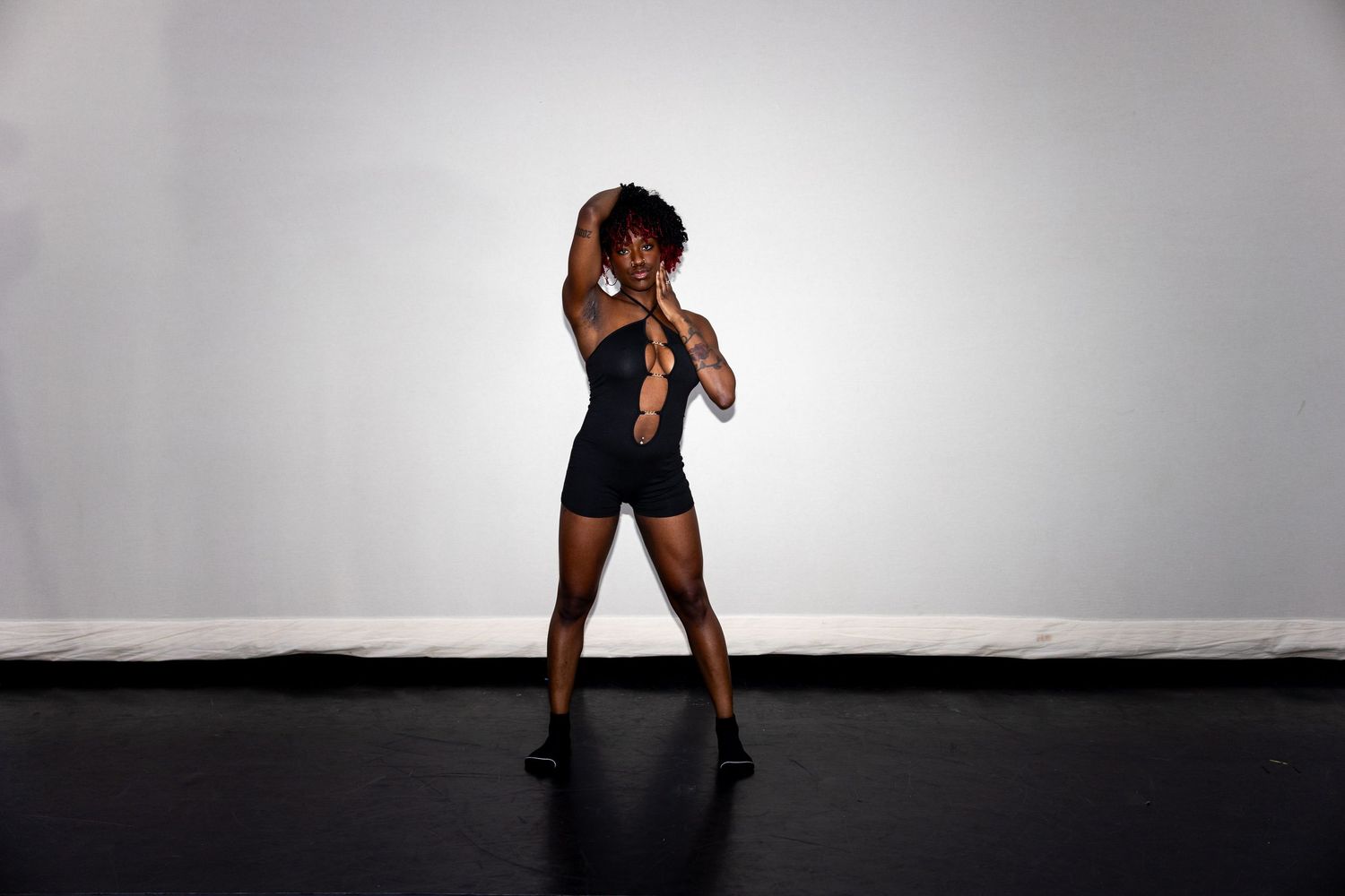 Woman in black shorts and top poses with arms raised in a minimalist studio setting.