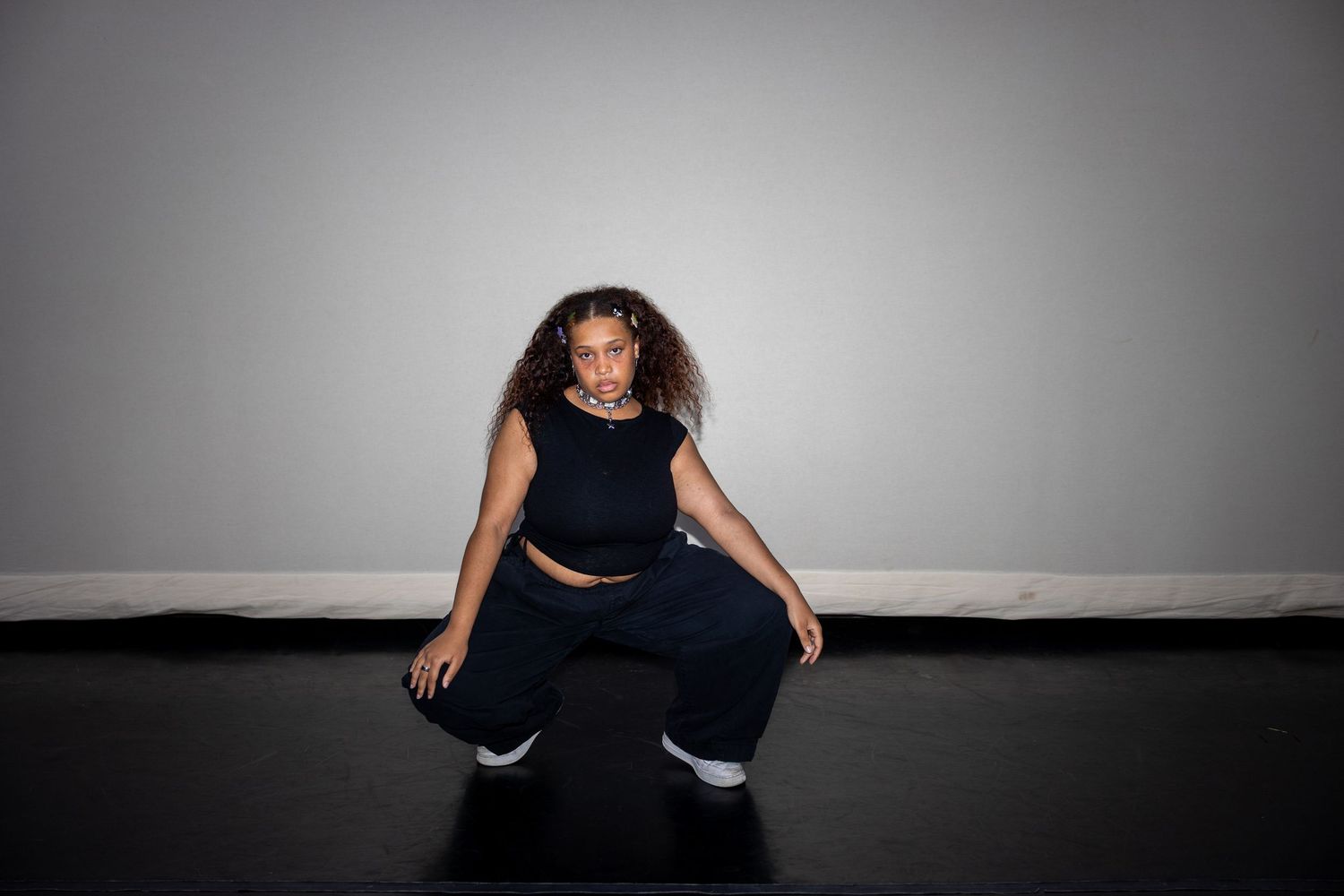 Young woman with curly hair in black outfit crouching in dynamic dance pose on black studio floor.