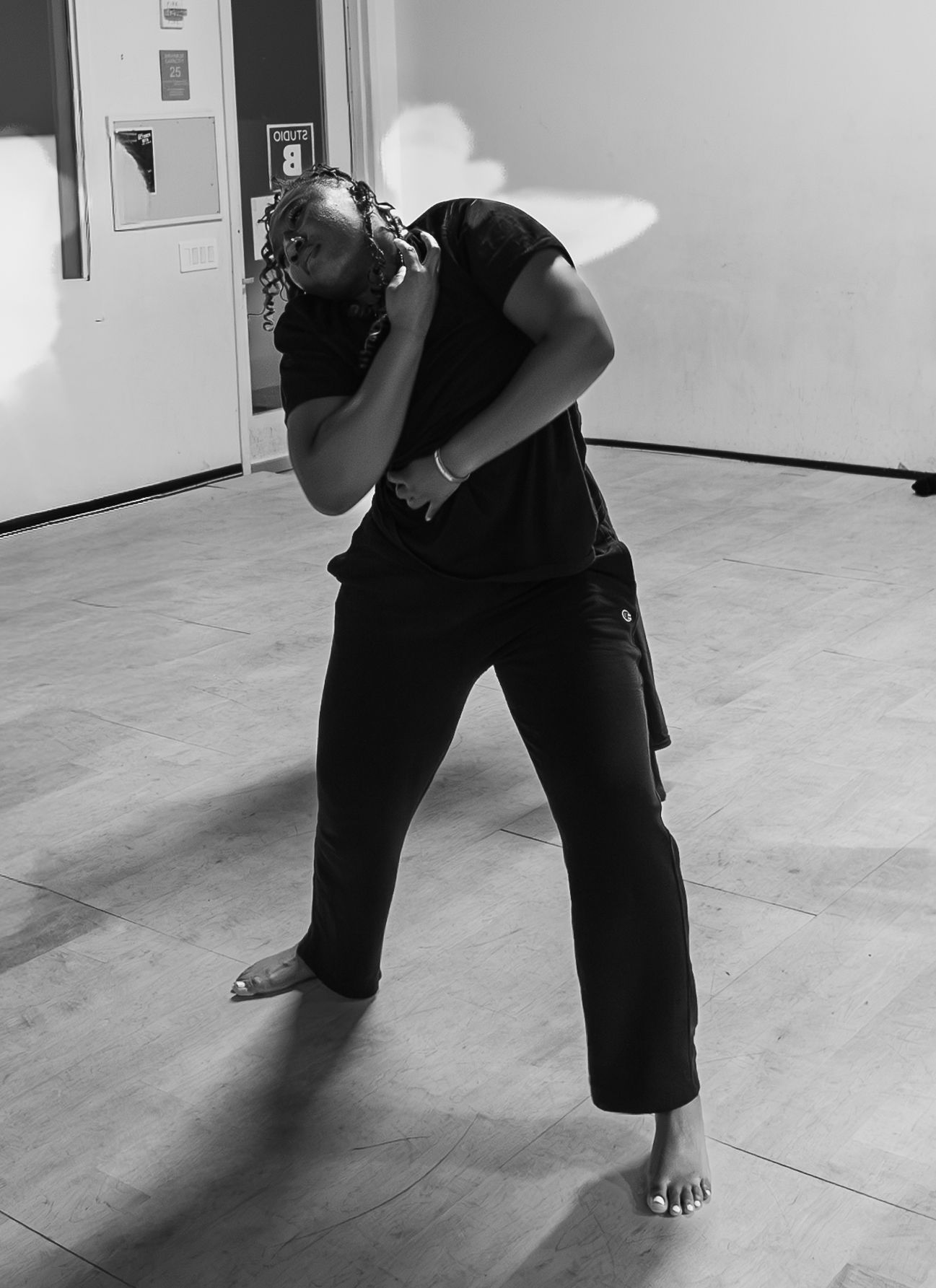 Black and white photo of a dancer in all-black outfit performing an expressive contemporary dance move barefoot indoors.