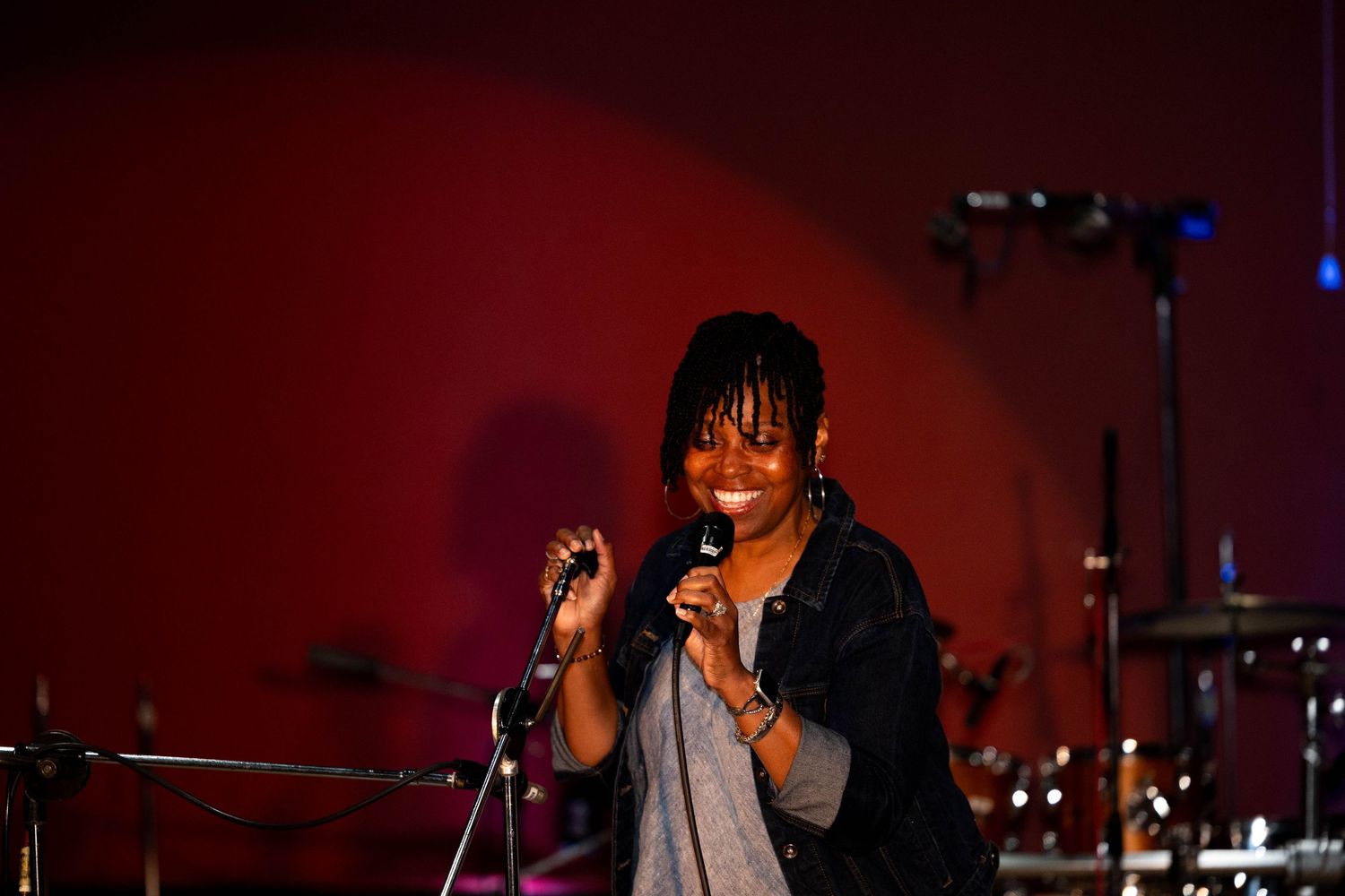 Smiling female vocalist performing on stage with microphone, wearing denim jacket, against red lit background.