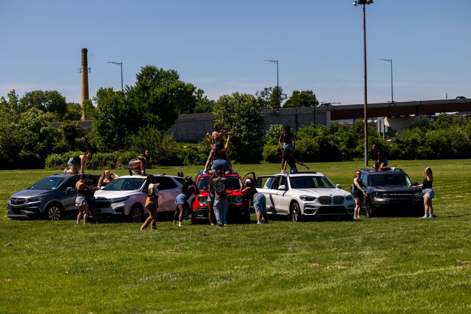 Group of people posing with multiple cars on a green grass field on a sunny day.