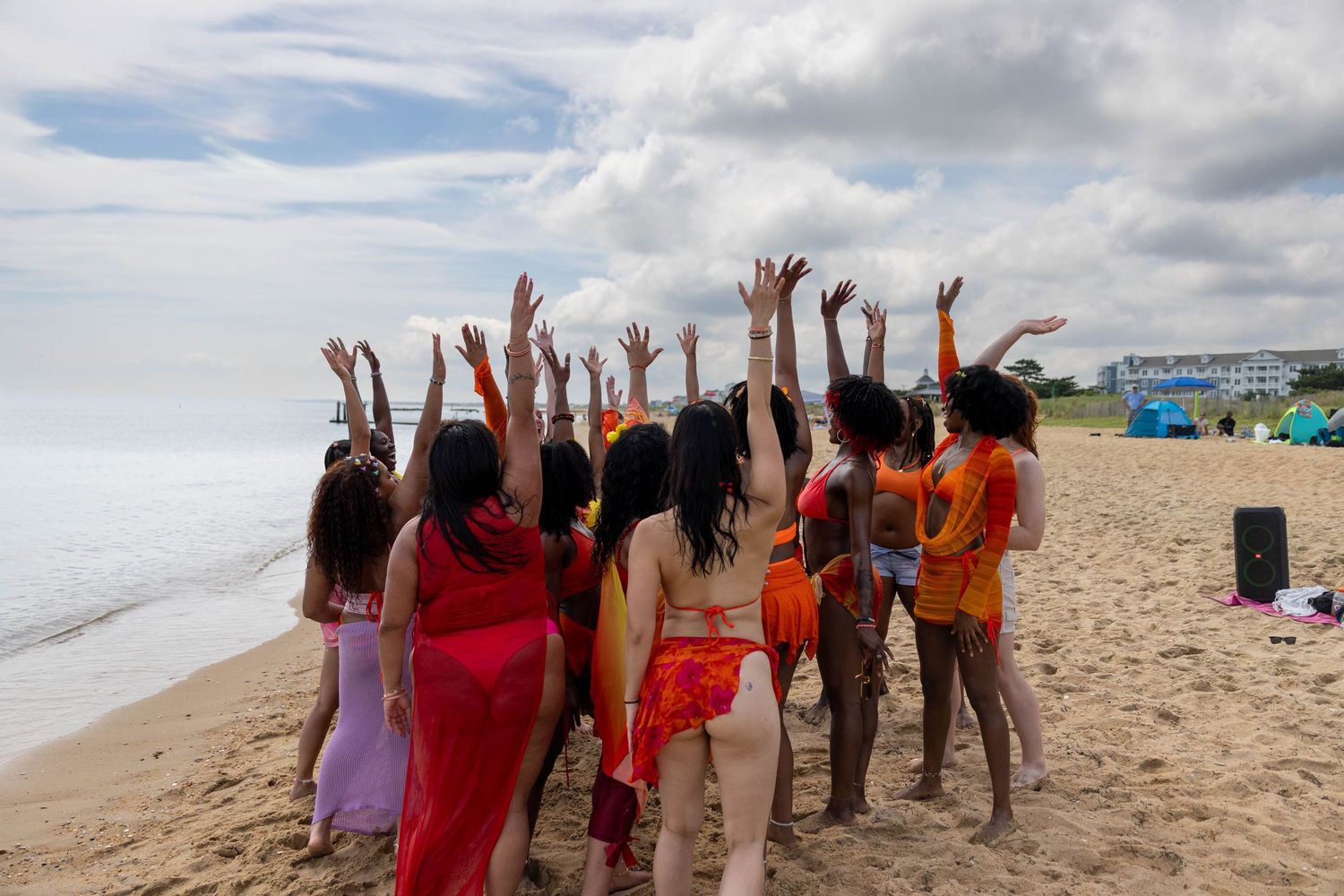 Group of women in colorful outfits raising hands on sandy beach with cloudy sky backdrop.
