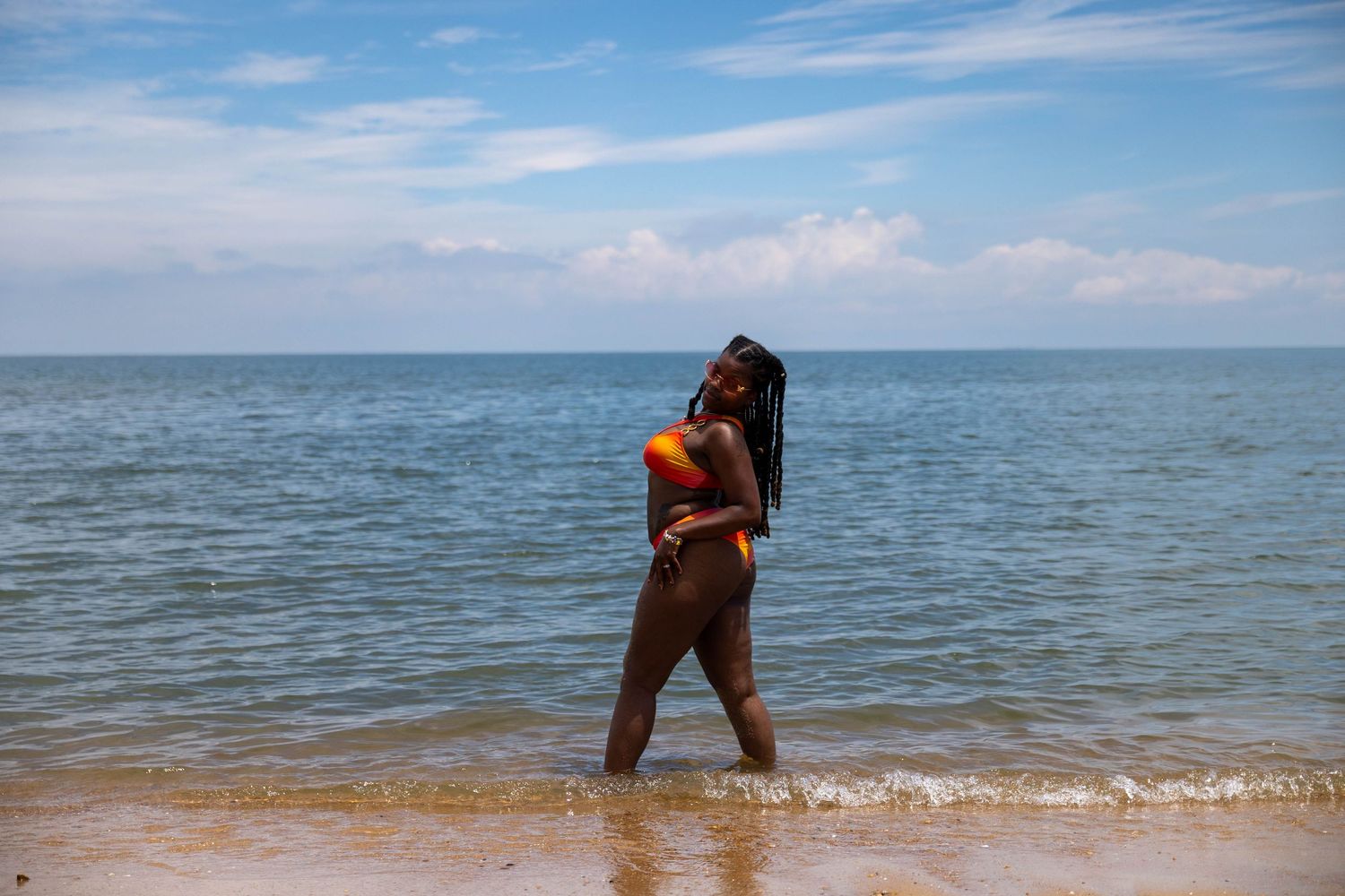 A woman in a colorful bikini stands in shallow ocean waves on a sunny beach with blue skies.