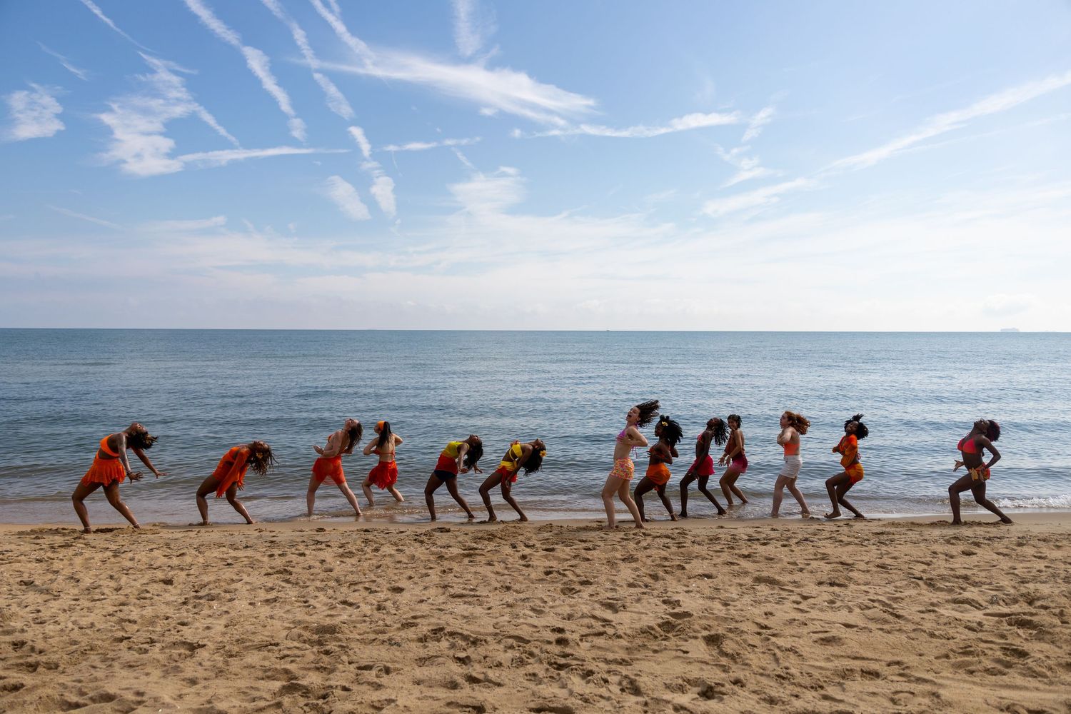 Group of people in orange shirts doing a beach workout exercise drill along the shoreline on a sunny day.