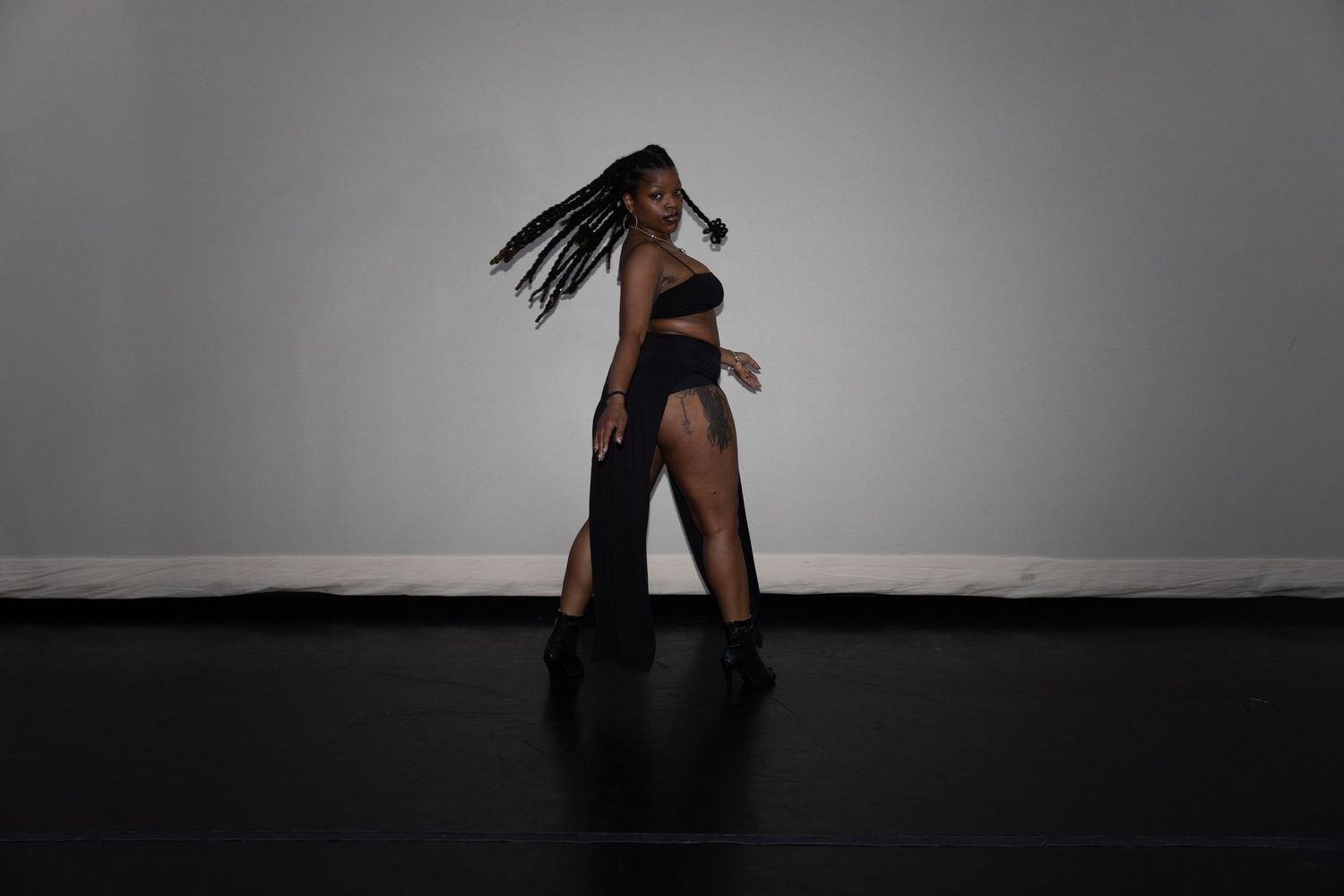 Woman in black outfit with braids dancing dynamically against gray studio background.