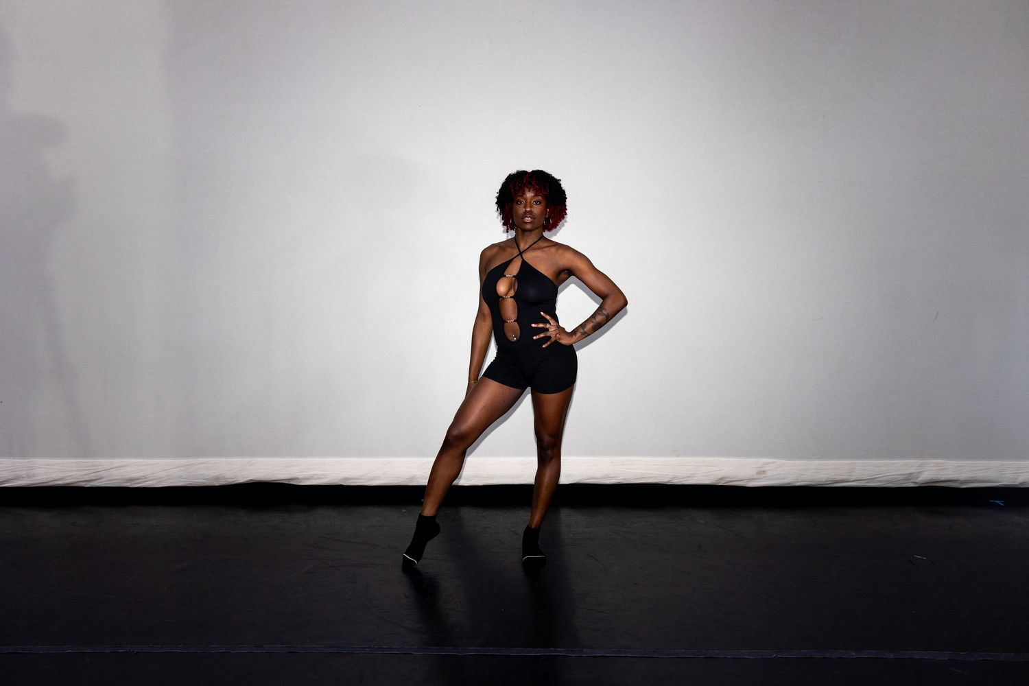 Black female dancer strikes elegant pose in black one-shoulder bodysuit on dark stage floor.