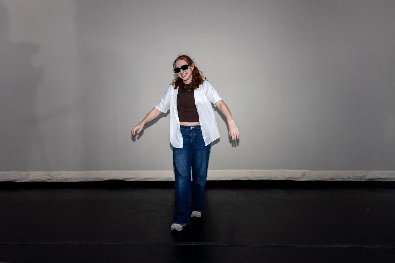 A dancer in jeans, white shirt, and sunglasses strikes a casual pose on a dark floor.