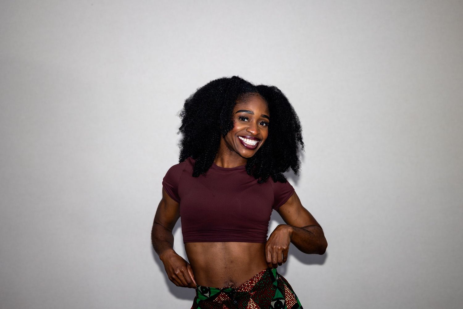 Smiling woman with natural afro in burgundy crop top and colorful skirt poses joyfully.