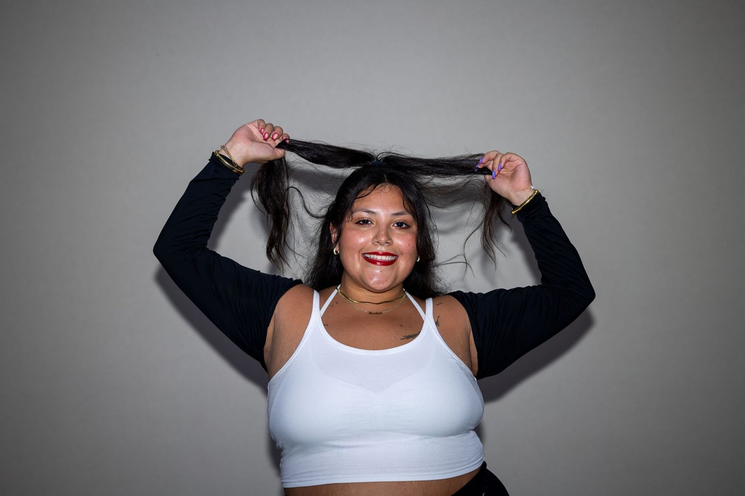 A plus-size woman in a white sports bra holds her long dark hair up while smiling against a gray background.
