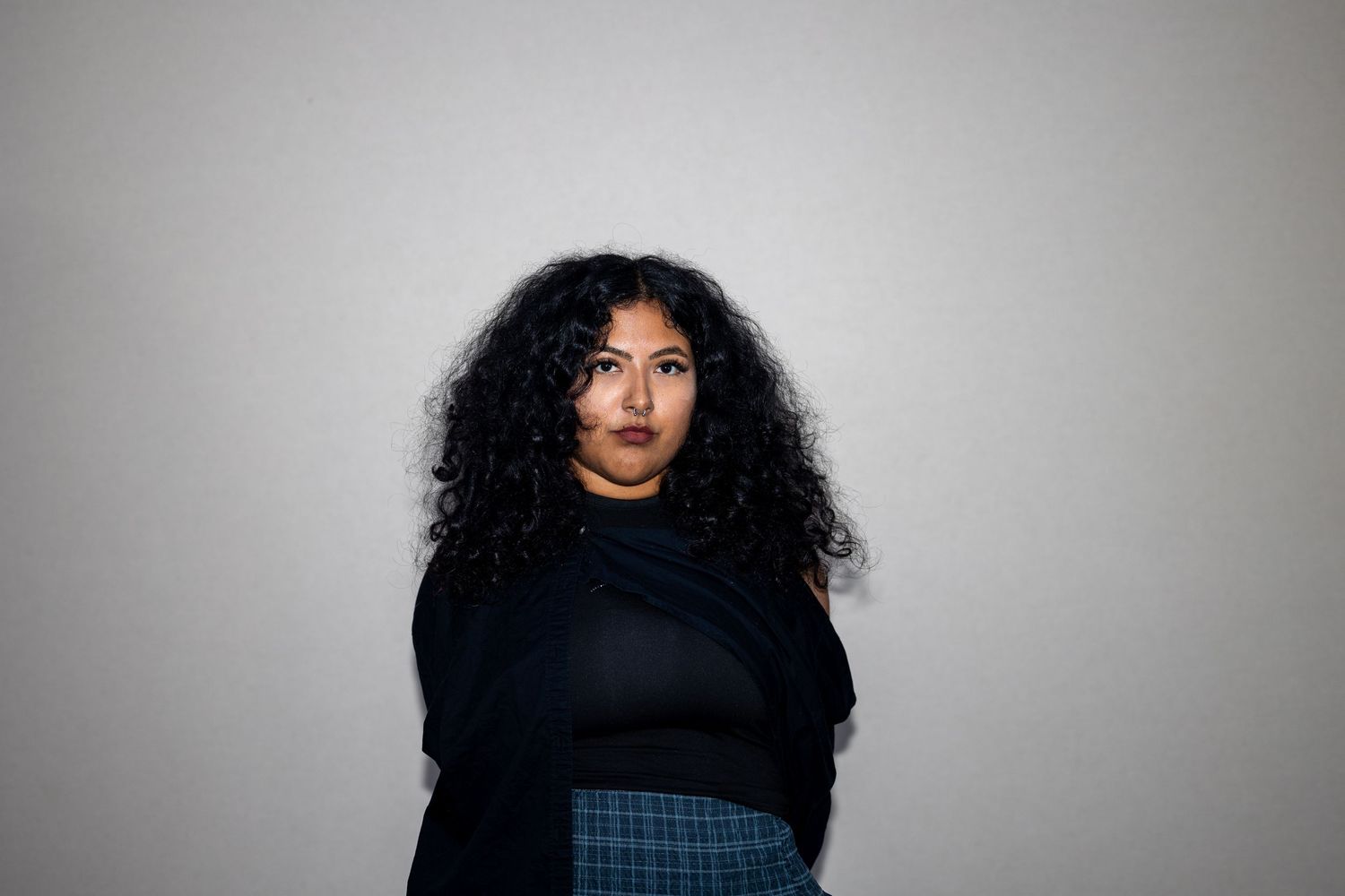 A woman with voluminous curly hair wearing a black turtleneck and plaid skirt poses against a gray background.
