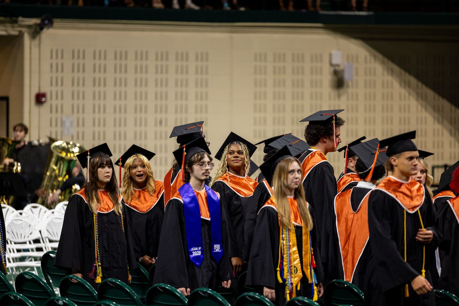 Graduates in black caps and orange gowns stand during a university commencement ceremony indoors.