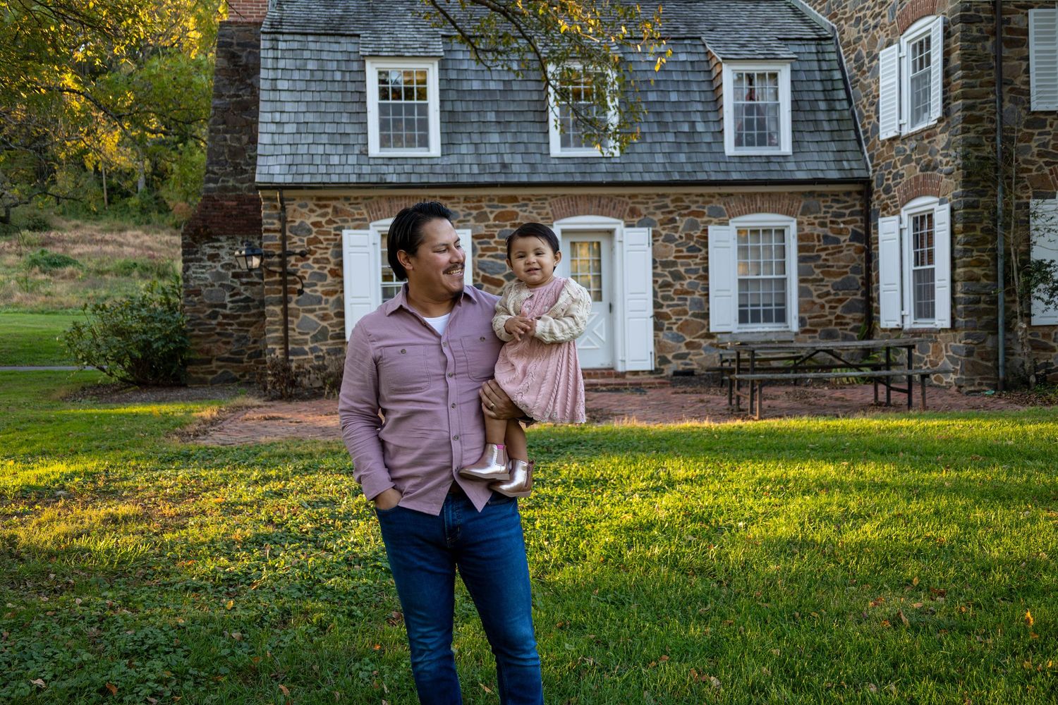 A man smiling and holding a young child on a green lawn in front of a historic stone house.