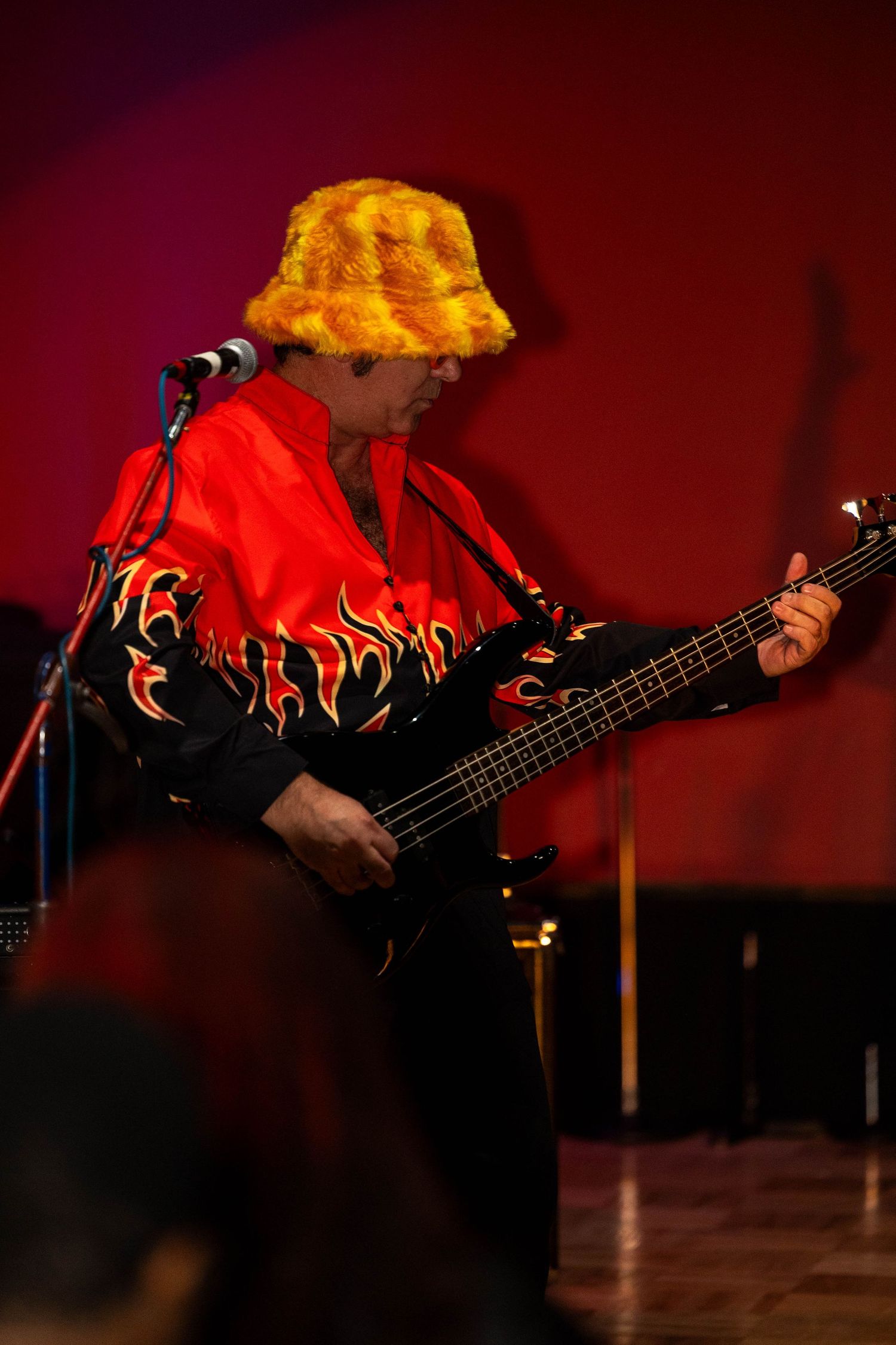 Musician in flame-print jacket and yellow bucket hat plays bass guitar on a red-lit stage.