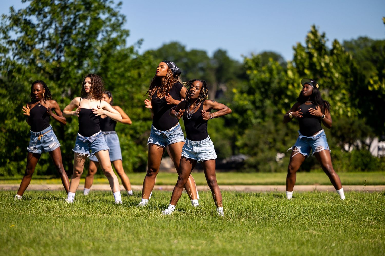 Group of young women dancing in sync outdoors on green grass with trees in background on a sunny day.